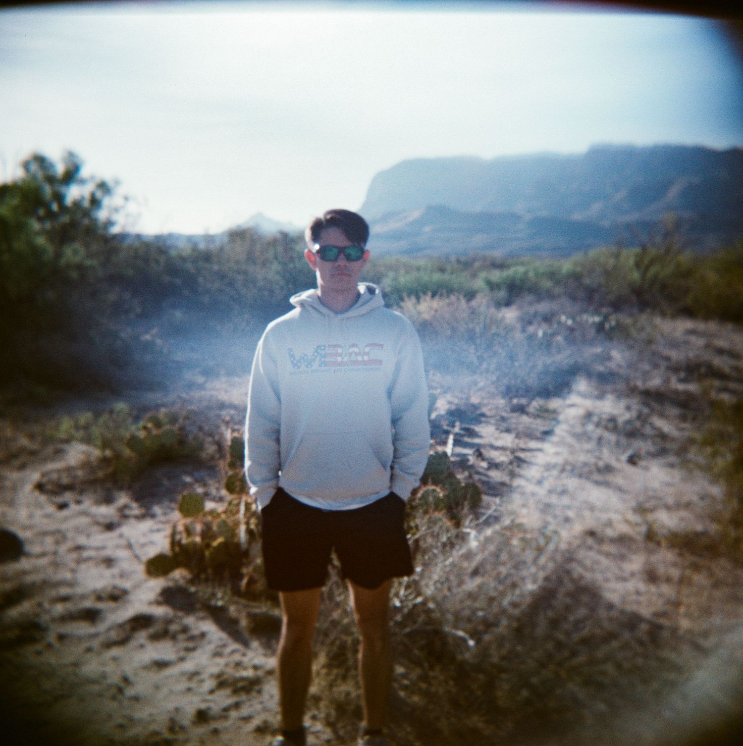 A young man in a white hoodie and sunglasses stands on a desert trail with rocky cliffs and shrubs in the background.