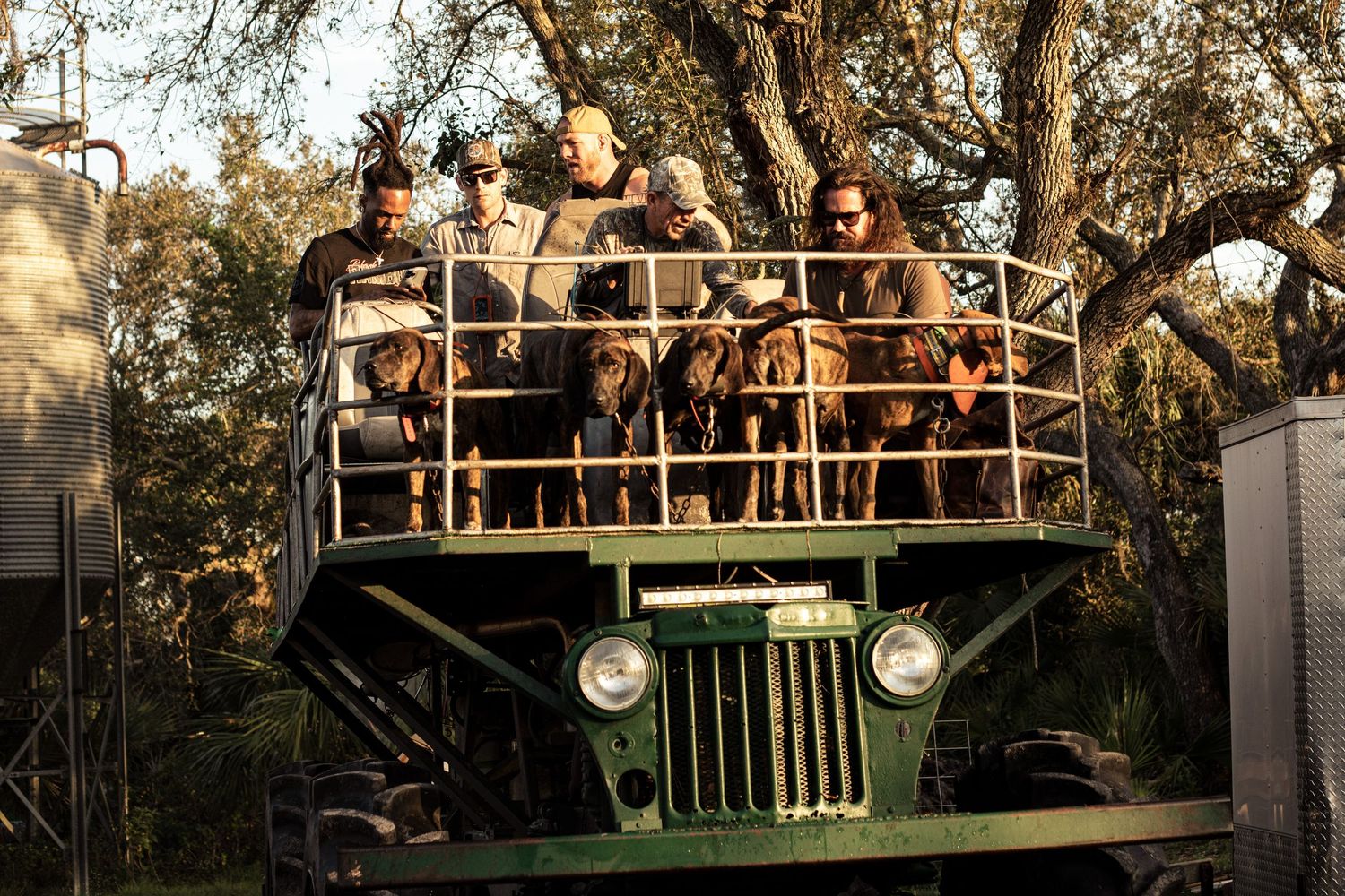 Group of people riding on a large green military vehicle through a wooded area on a sunny day.