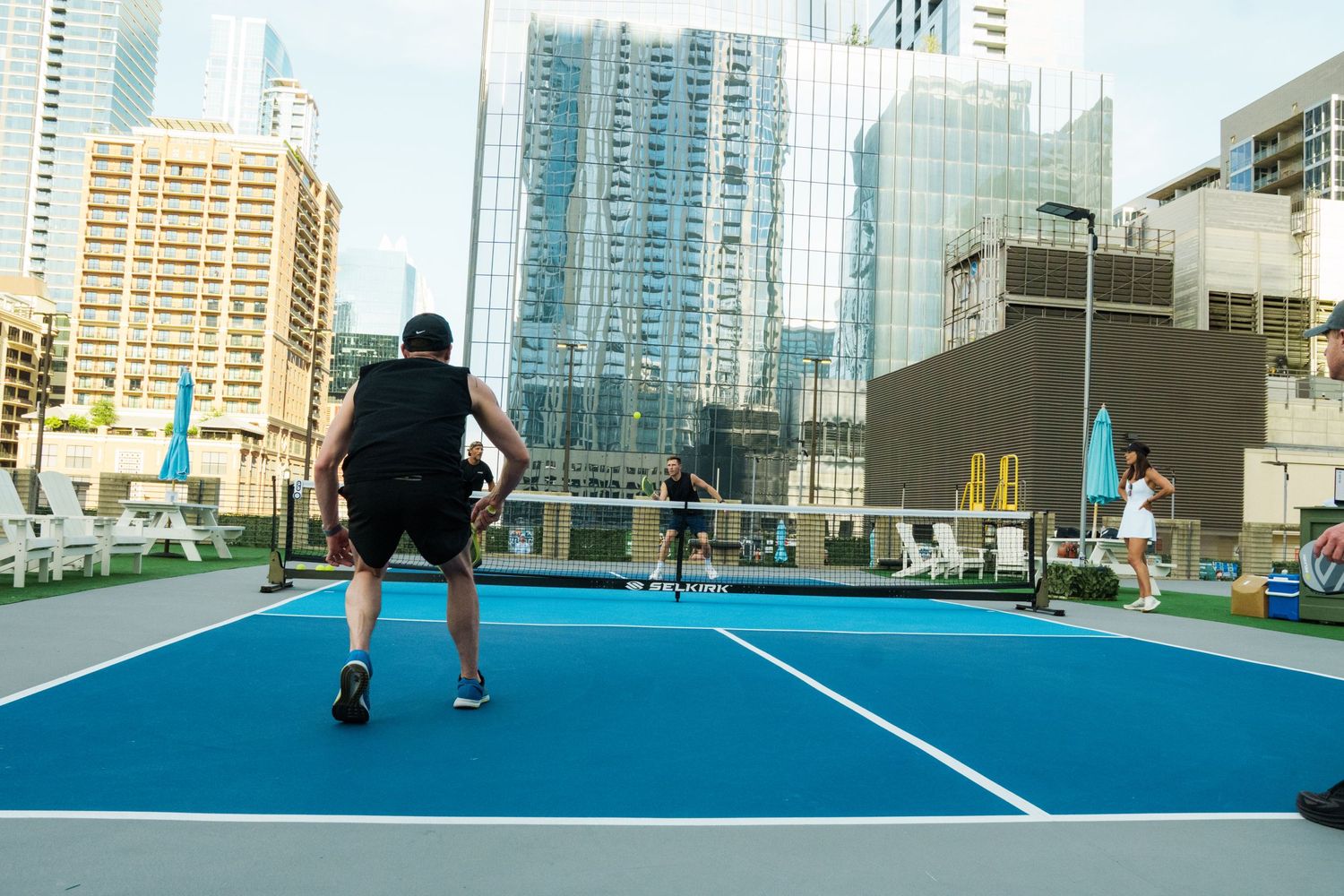 Players compete on a blue rooftop pickleball court surrounded by urban skyscrapers in a modern city setting.