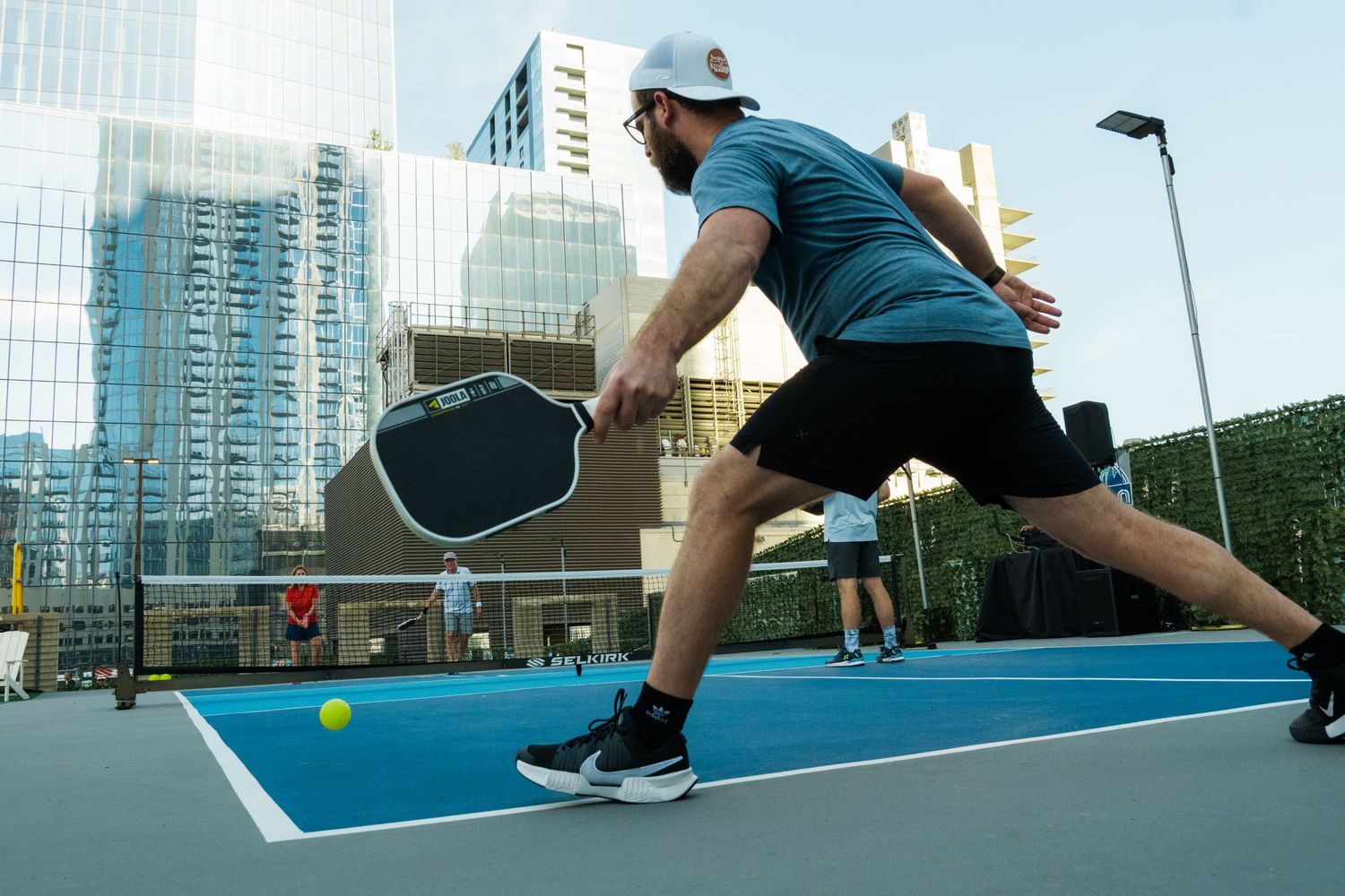 Man playing pickleball on rooftop court in urban city setting, lunging to hit yellow ball with paddle.