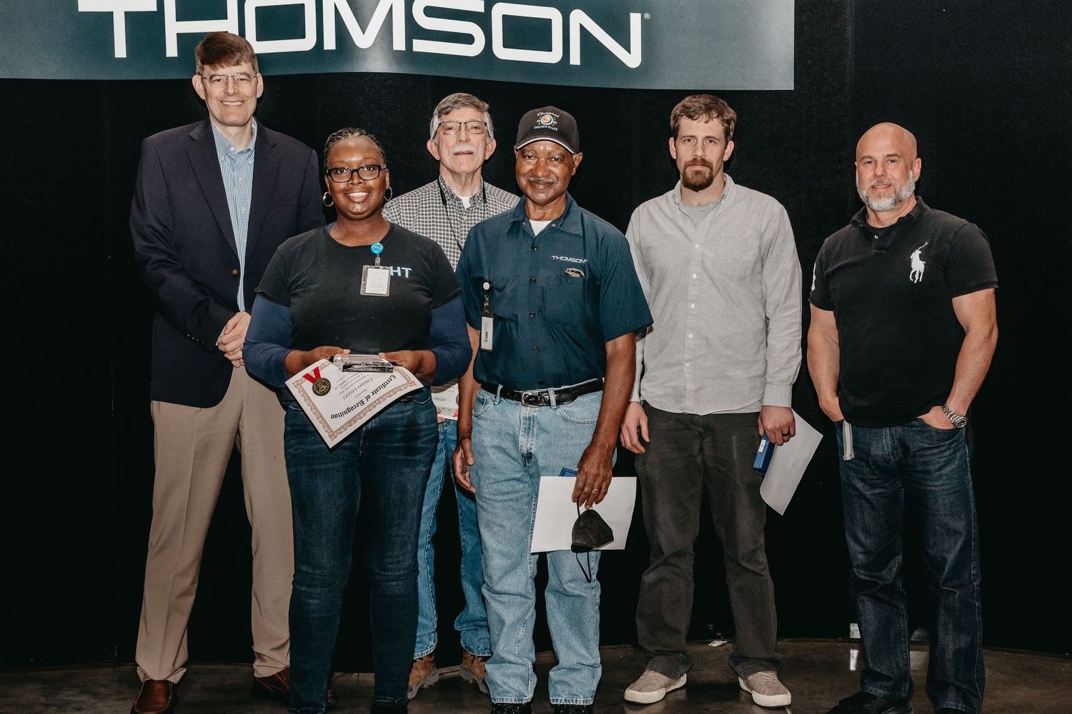 Group of six professionals posing together in front of a Thomson sign at a corporate recognition event.