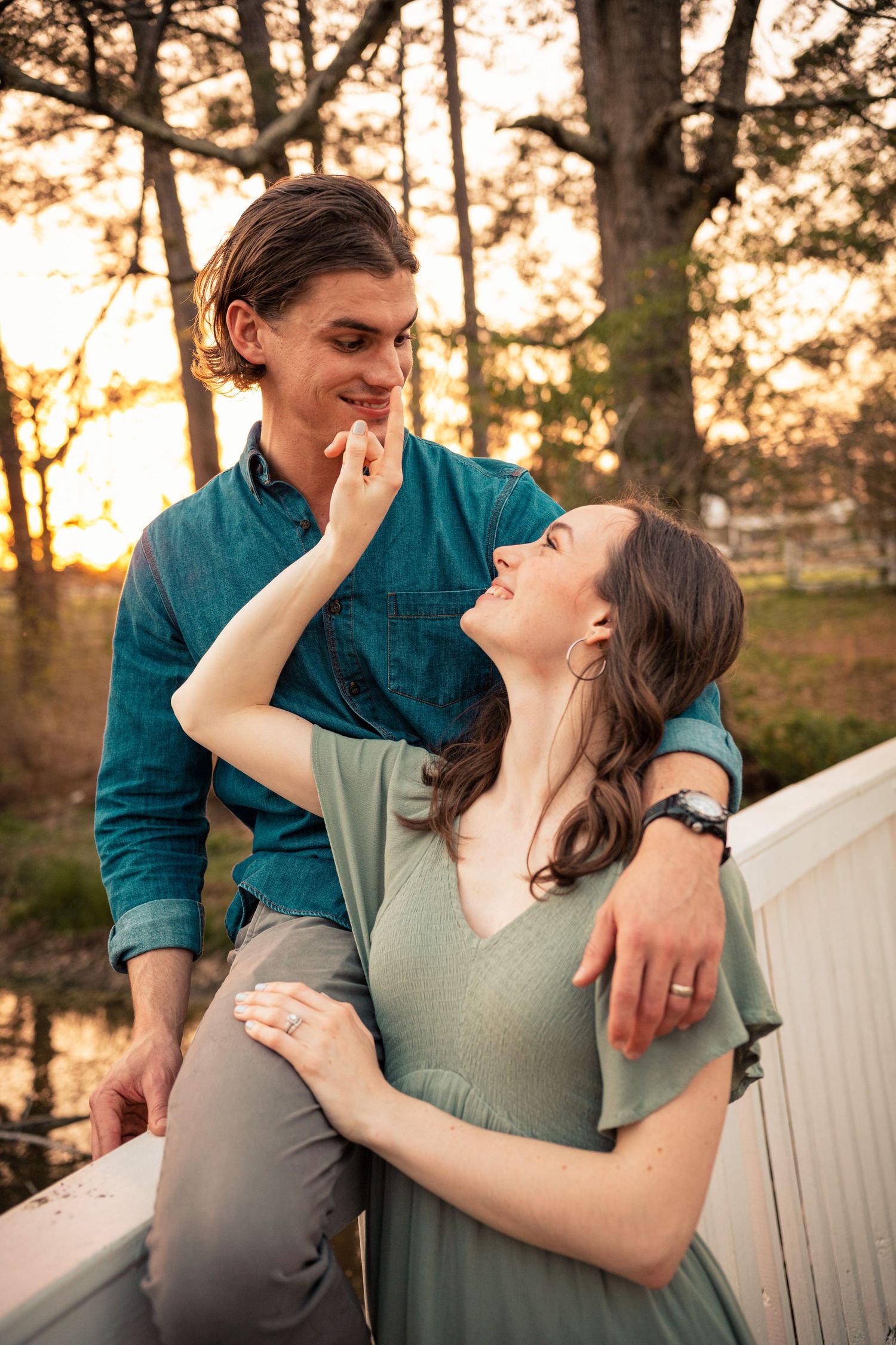 Couple sharing a romantic moment at sunset, woman in sage dress touching man's face near a white fence.