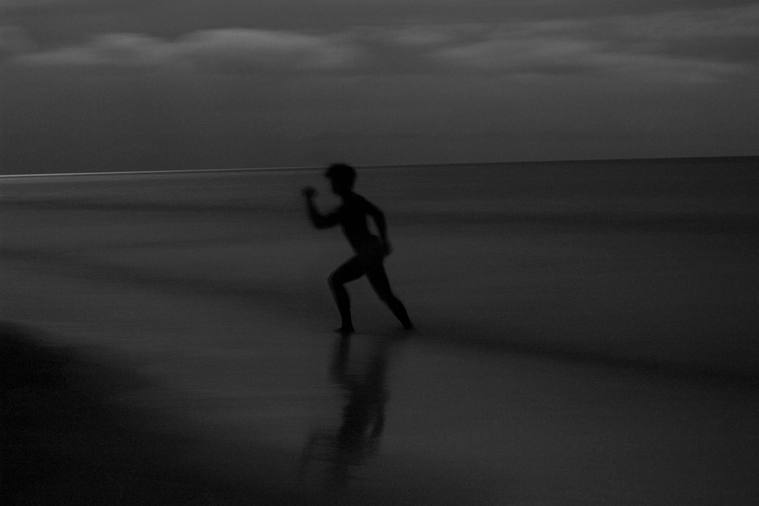 Silhouette of a person running along a dark beach at dusk in a moody black and white long exposure photo.