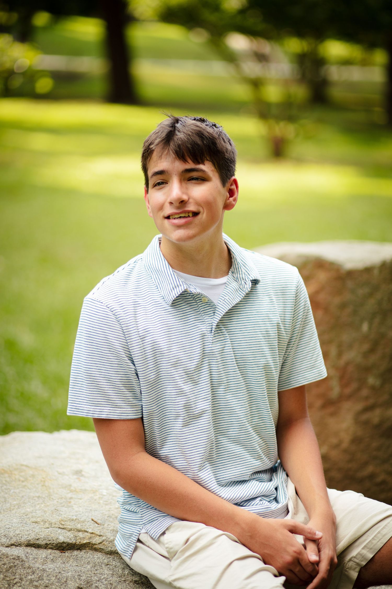 Smiling teenage boy in striped polo sitting on large rock in green park.