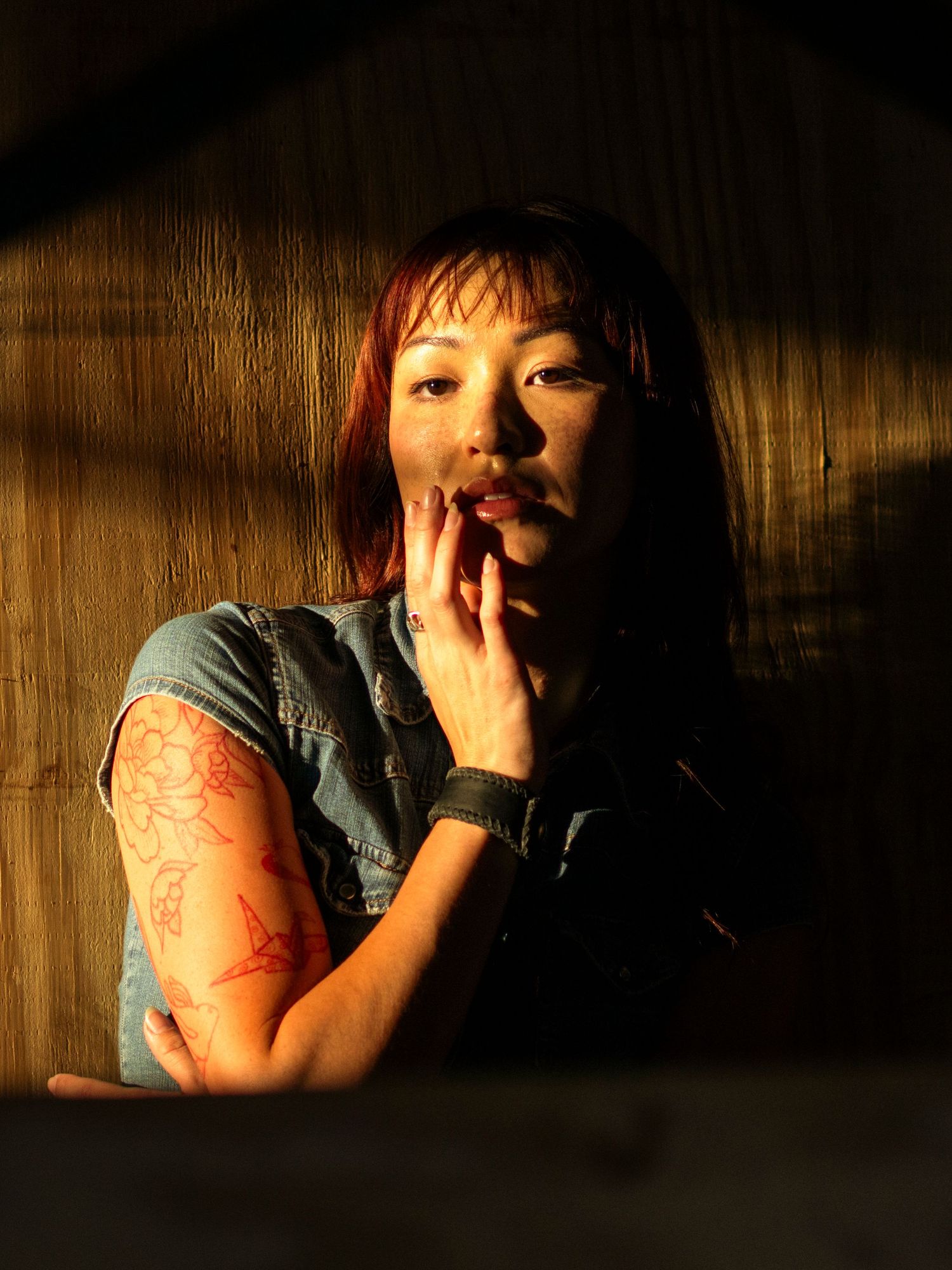 Young Asian woman with red tattoos on her arm in dramatic sunlight against a wooden wall.