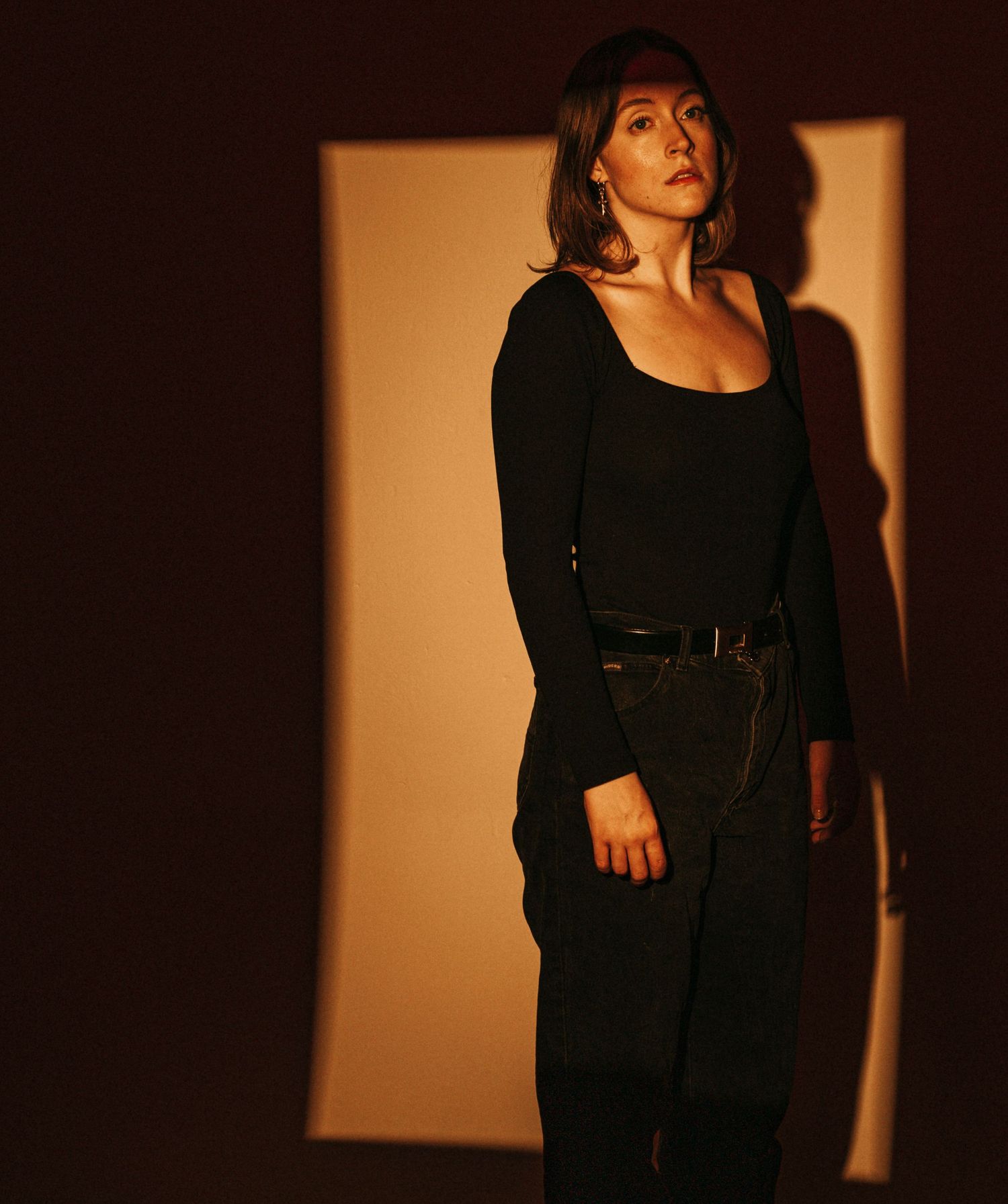 Woman in black bodysuit and dark pants stands in dramatic warm shadow lighting.