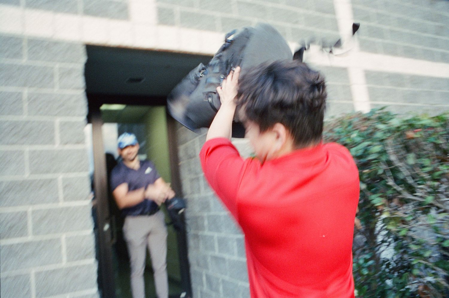 Person in red shirt raising bag over head near brick building entrance.