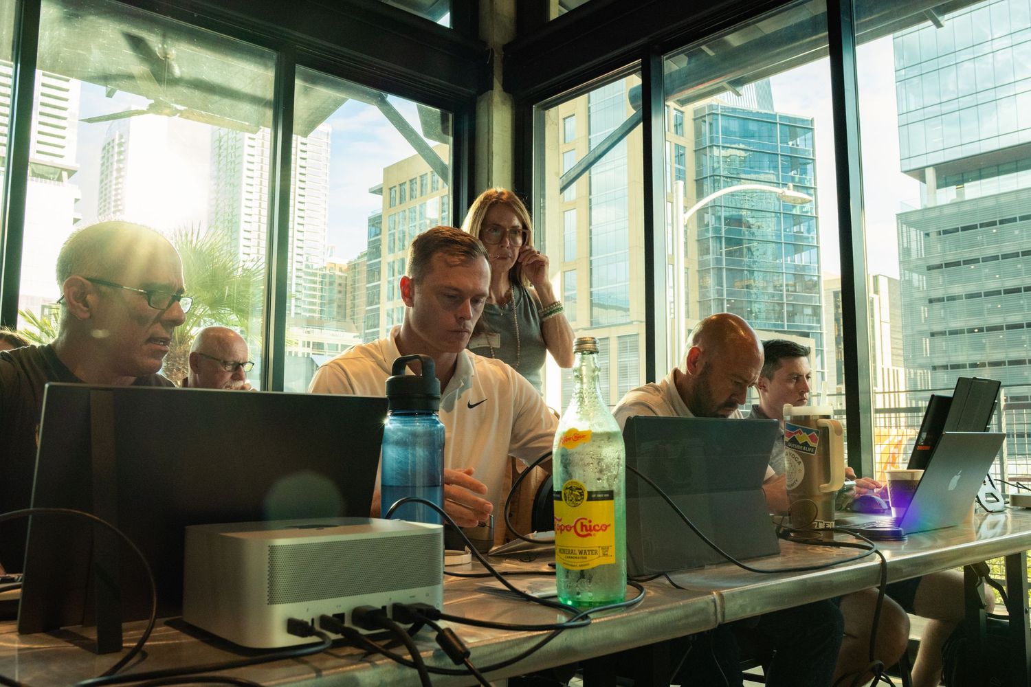 Professionals working on laptops in a modern glass office with city skyline views in bright sunlight.