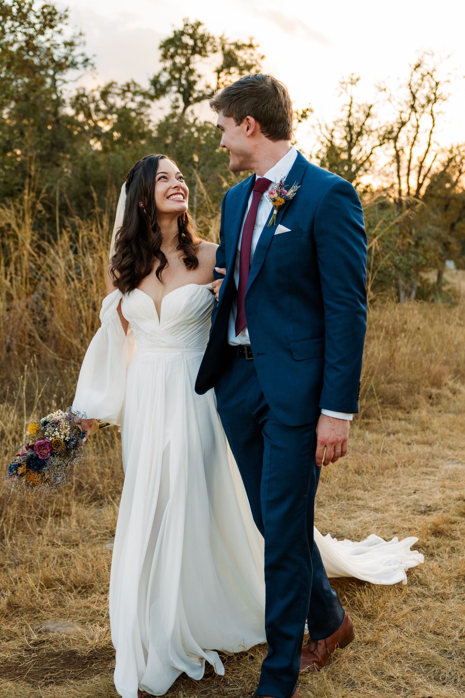 Bride in white gown and groom in navy suit share a joyful moment outdoors at golden hour.