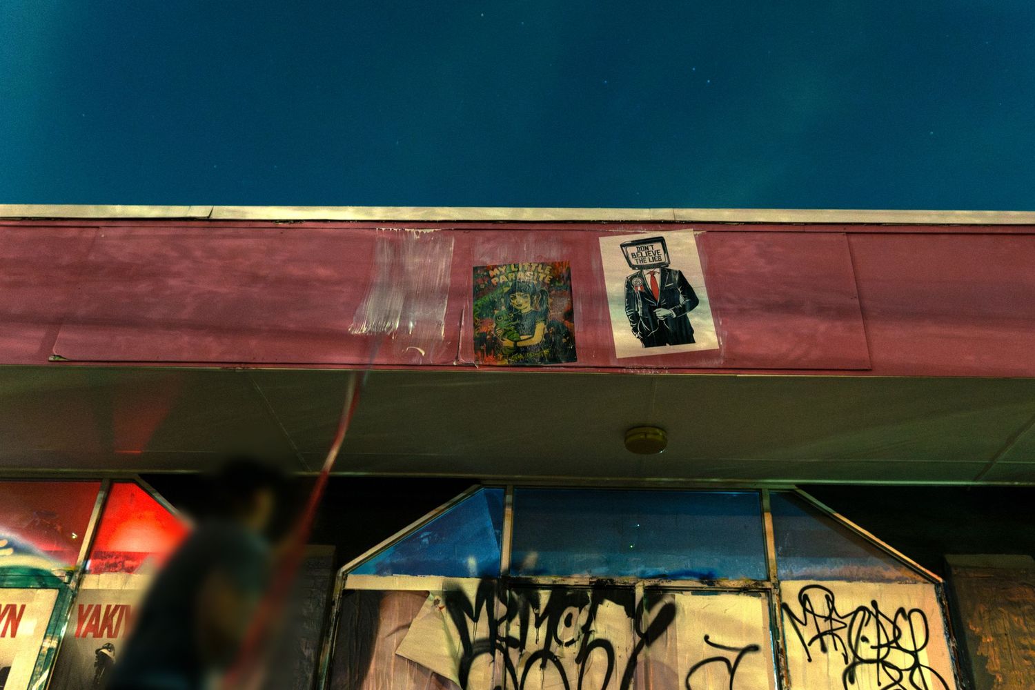 Graffiti-covered storefront at night with posters on pink facade under deep blue sky.