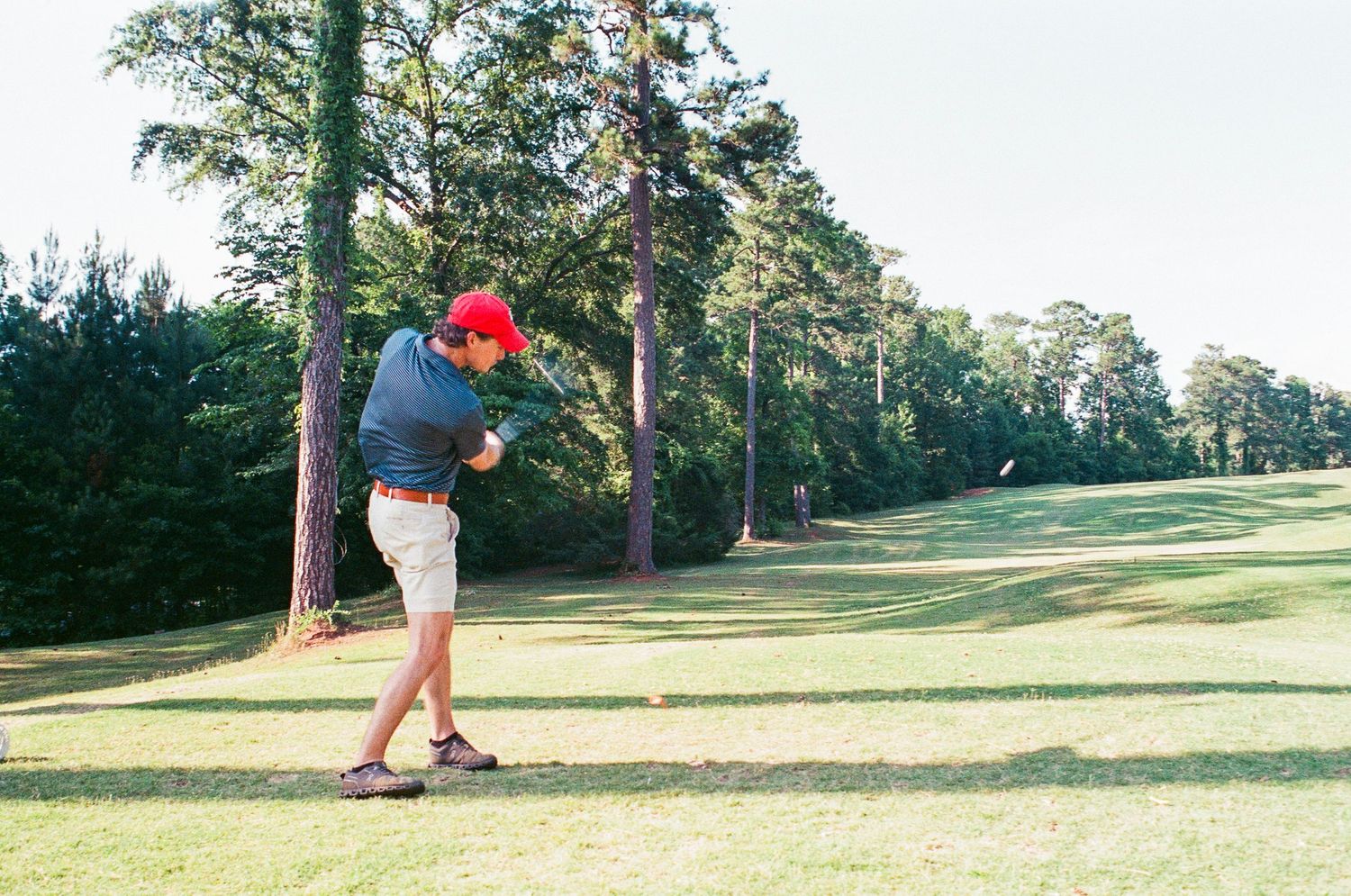 Golfer in red cap and khaki shorts swinging a golf club on a sunny tree-lined fairway.