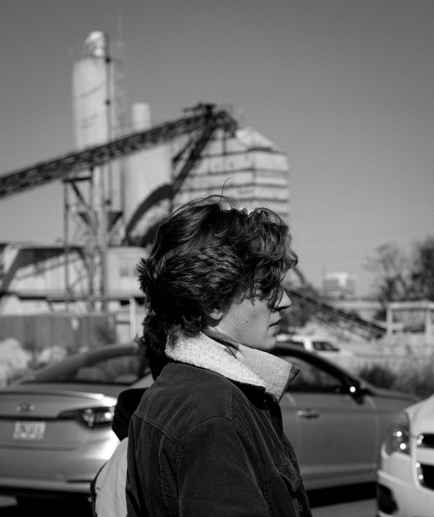 Black and white photo of a young man with curly hair standing outdoors near parked cars and industrial structures.