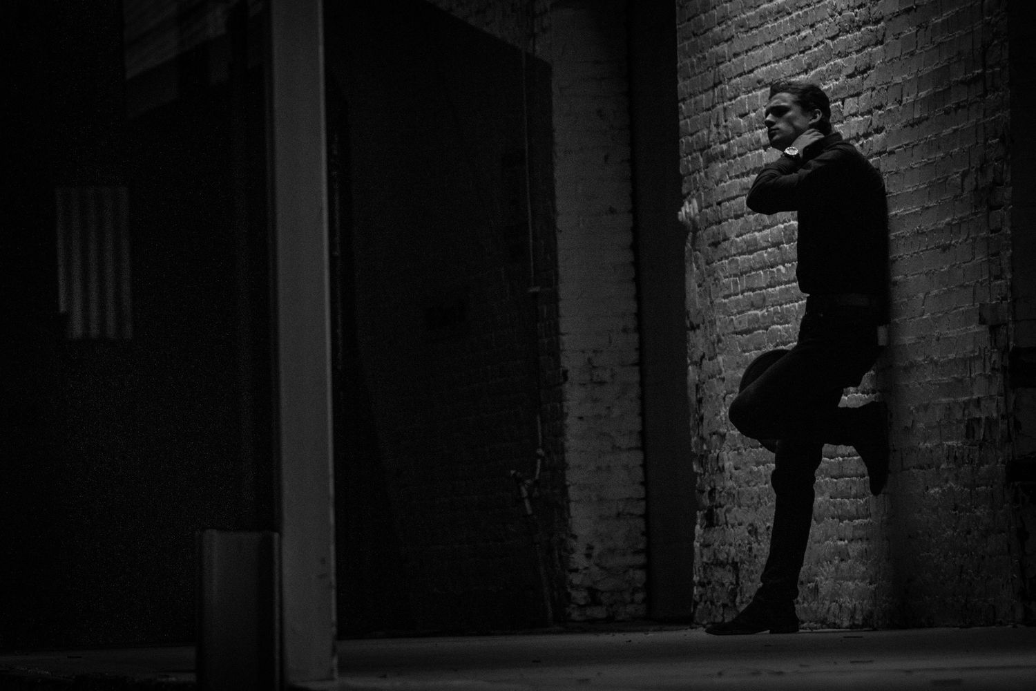Man in black suit leaning against brick wall in dramatic black and white photography.