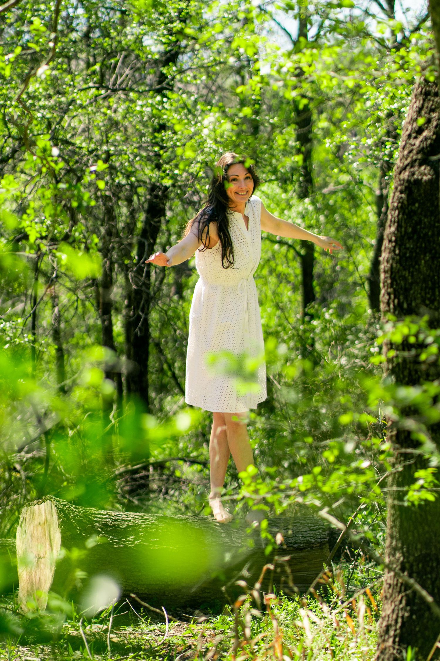 Joyful woman in a white dress balances on a fallen log with arms outstretched, surrounded by vibrant green forest bathed in sunlight.