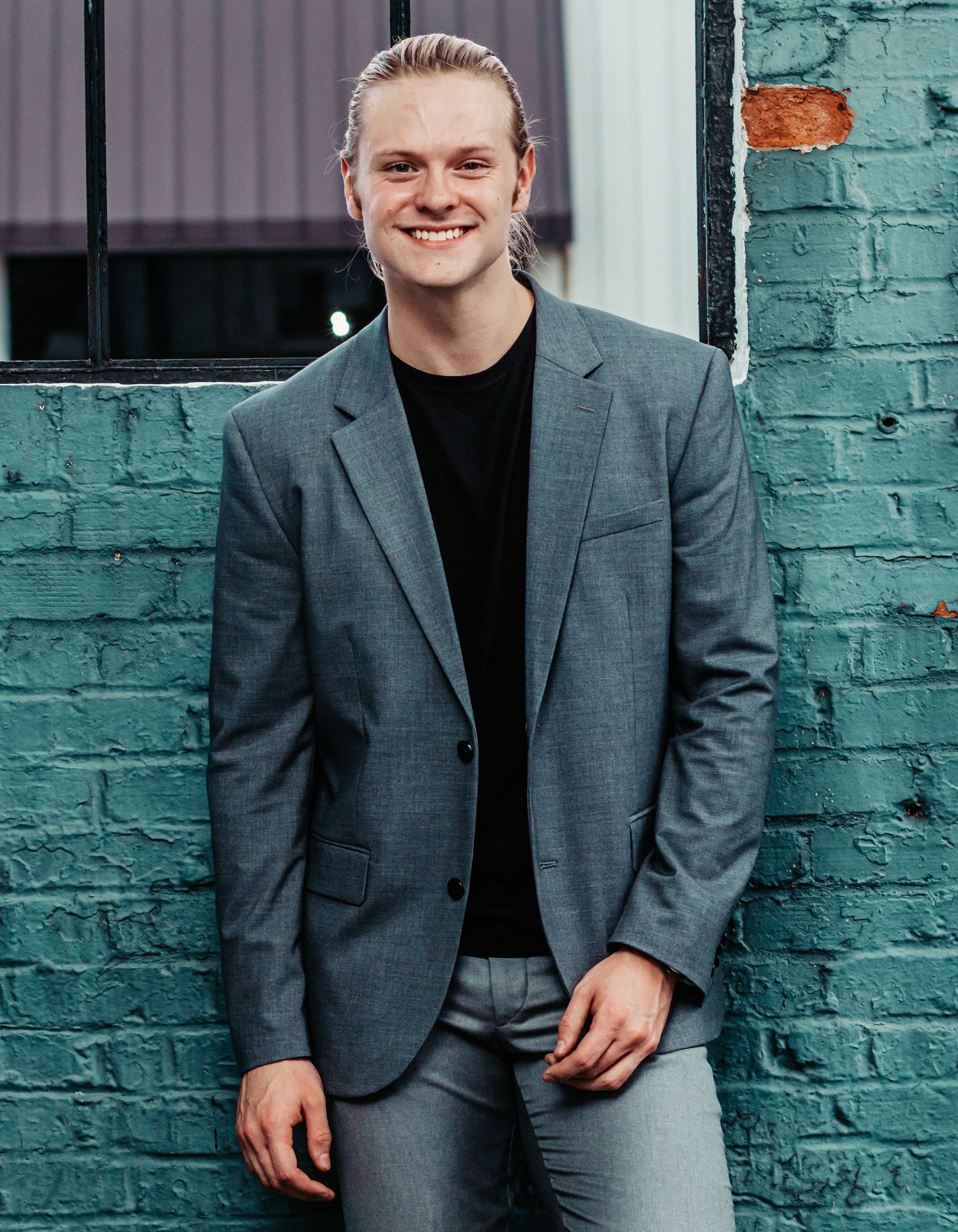 Young professional man in gray blazer smiling against teal brick wall outdoors.
