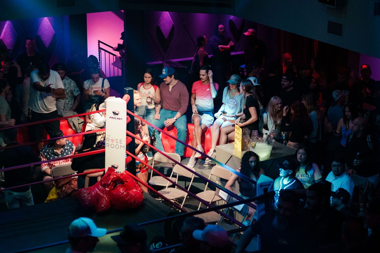 Overhead view of colorful wrestling ring with performers and seated audience in a vibrant venue.