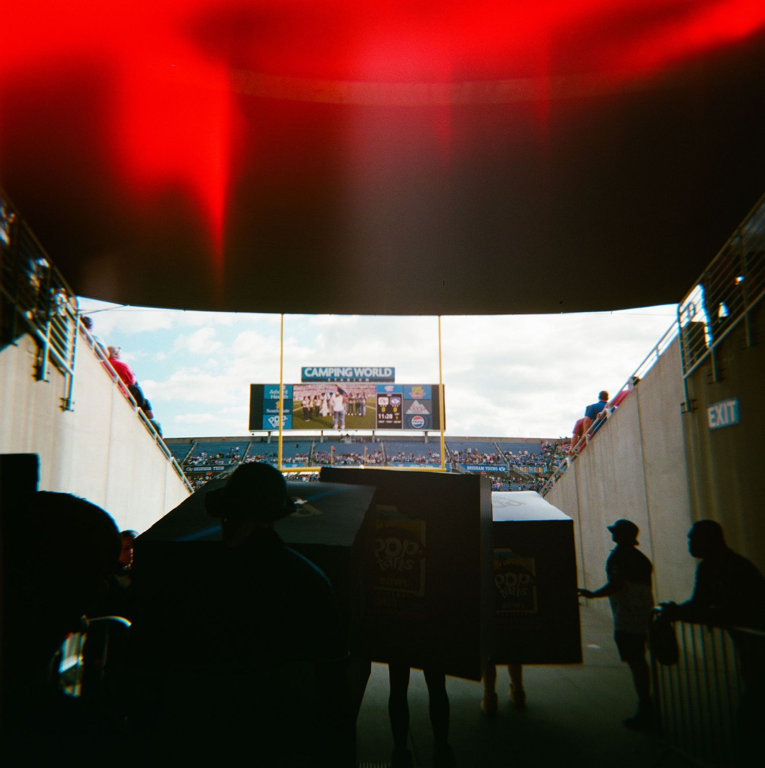 Silhouetted figures walk through a dark stadium tunnel toward a bright football field with a scoreboard.