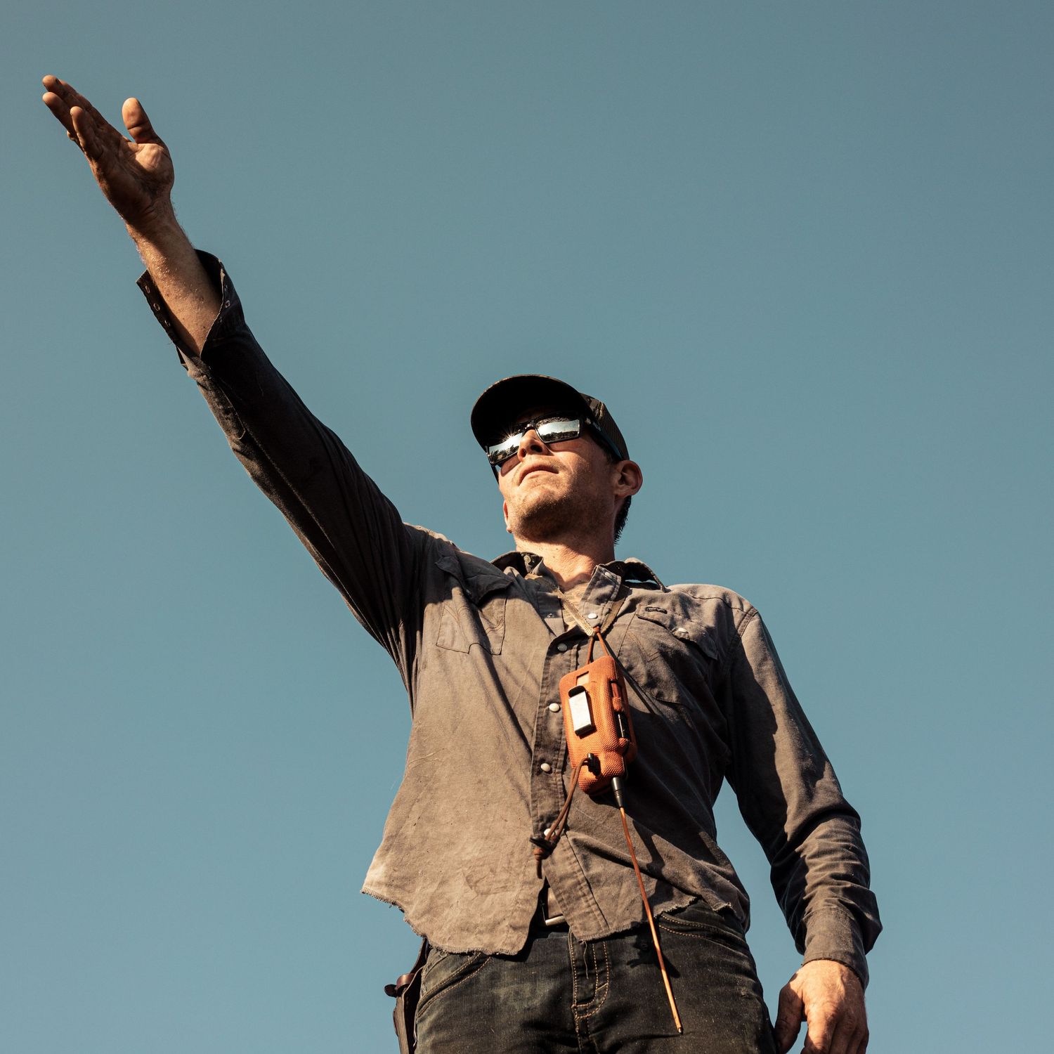 A man in a cap and sunglasses raises one arm toward the sky while holding a camera against a clear blue sky.
