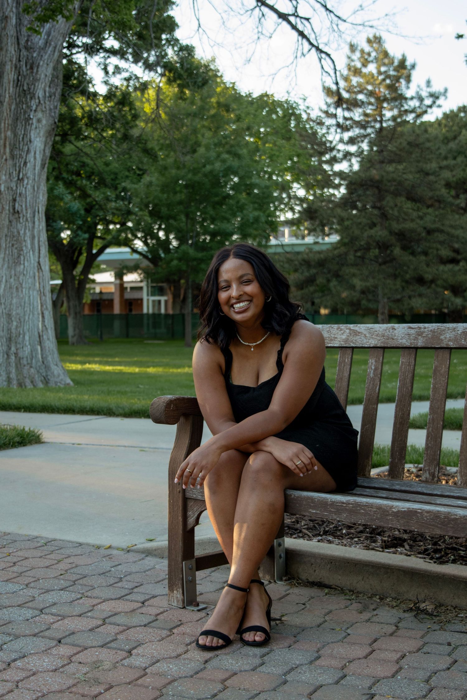 A smiling young woman in a black dress sits on a wooden bench in a lush green park on a warm day.