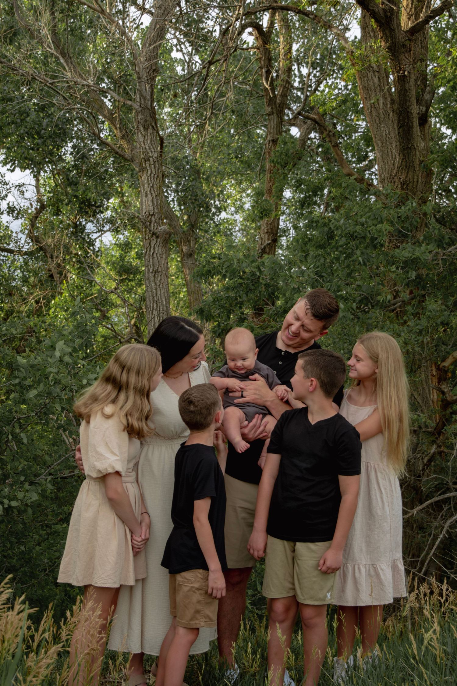 Large family with parents and five children gathering outdoors among lush green trees during a summer portrait session.