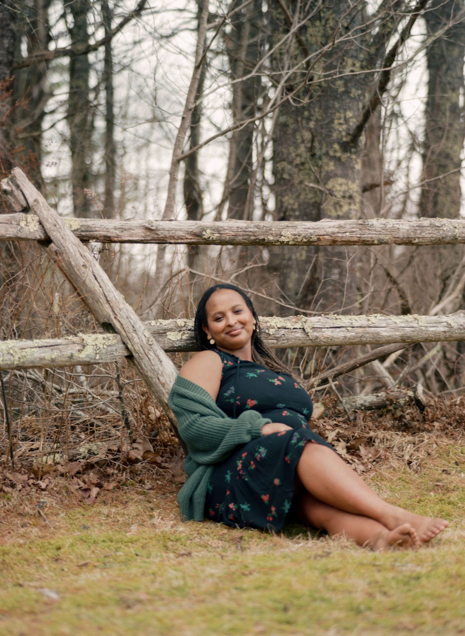 Pregnant woman in floral dress and green cardigan sitting barefoot against rustic wooden fence in wooded outdoor setting.