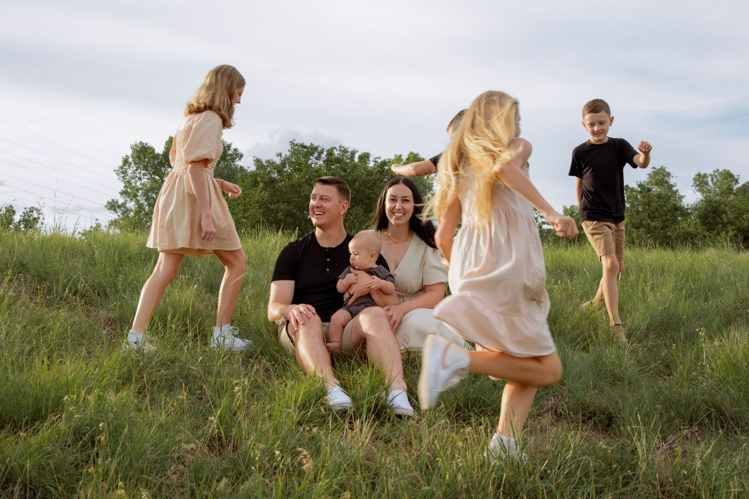Family enjoying outdoor time in a green field, with parents sitting on grass while children play around them on a cloudy day.