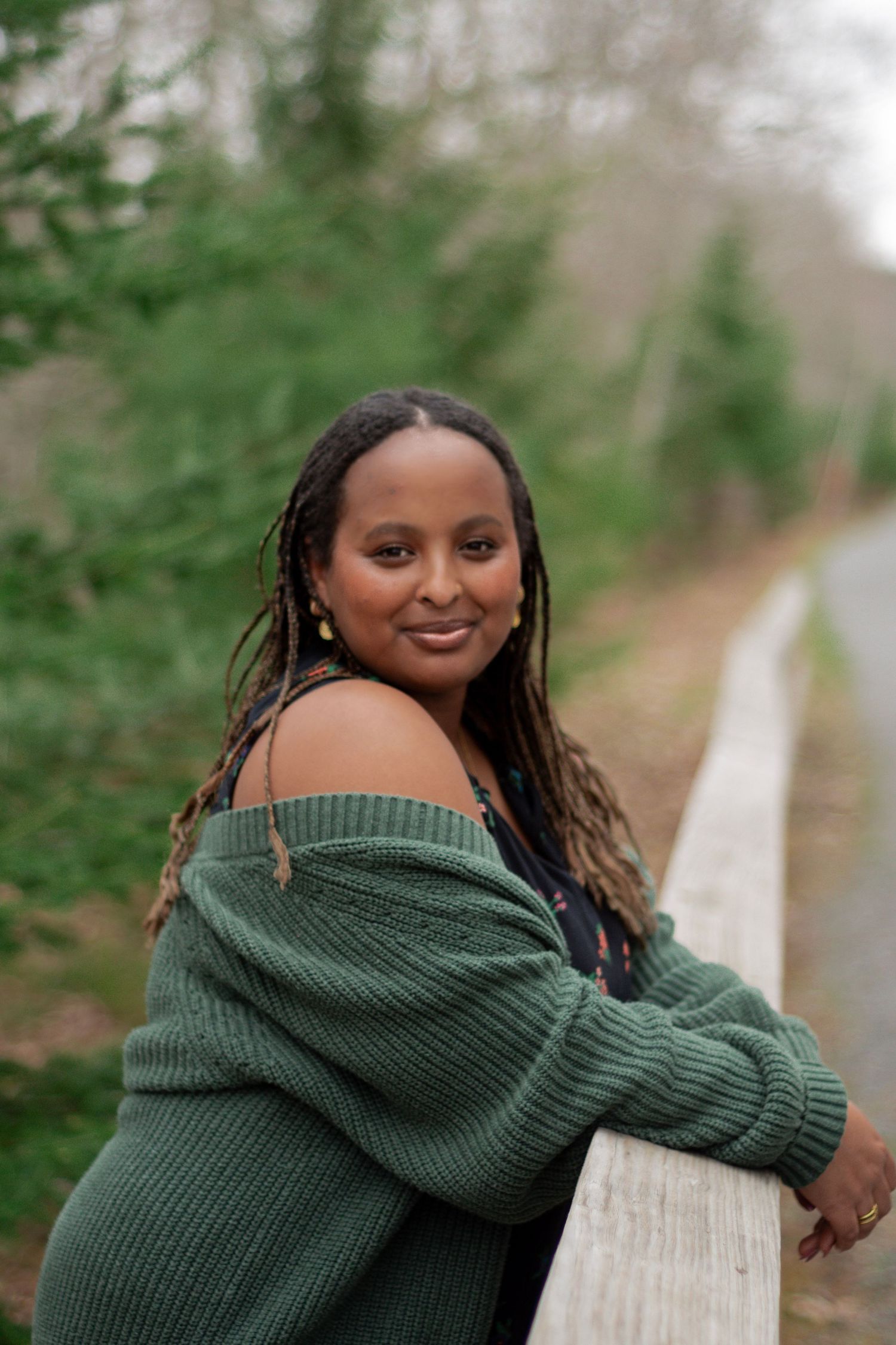 Woman in green off-shoulder sweater smiling outdoors, leaning on white fence with evergreen trees in background.
