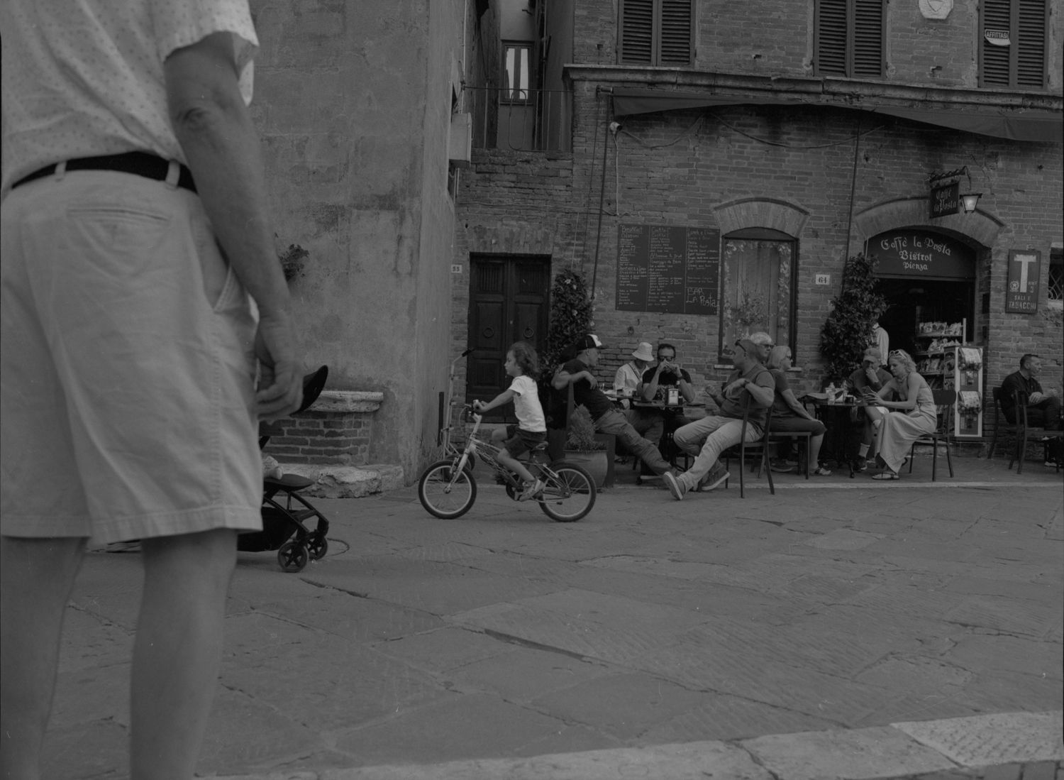 Black and white street photo of people relaxing at outdoor cafe while a child rides a bicycle in a town square.