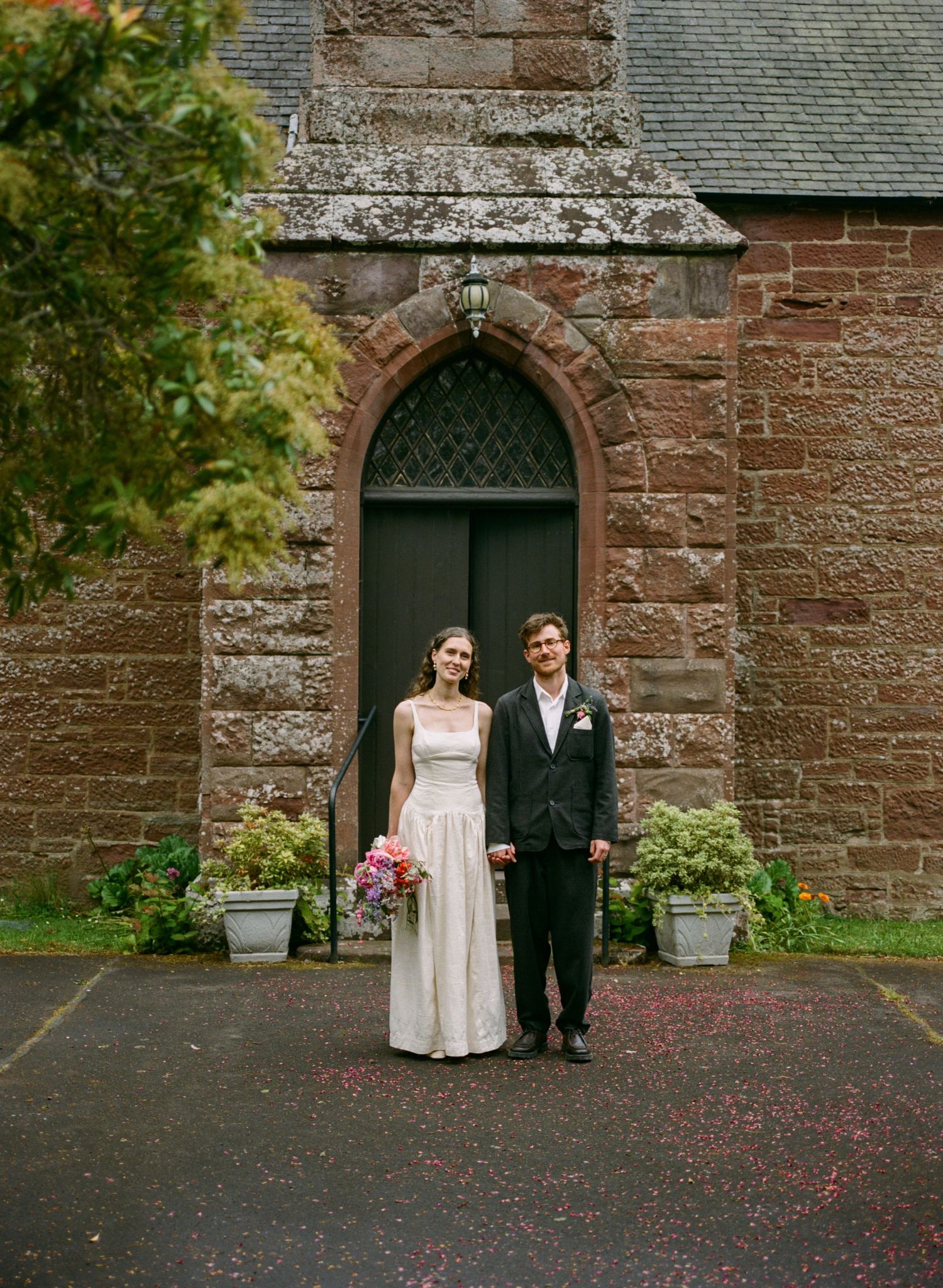 Bride and groom stand holding hands before a stone church door with rose petal confetti on the ground.