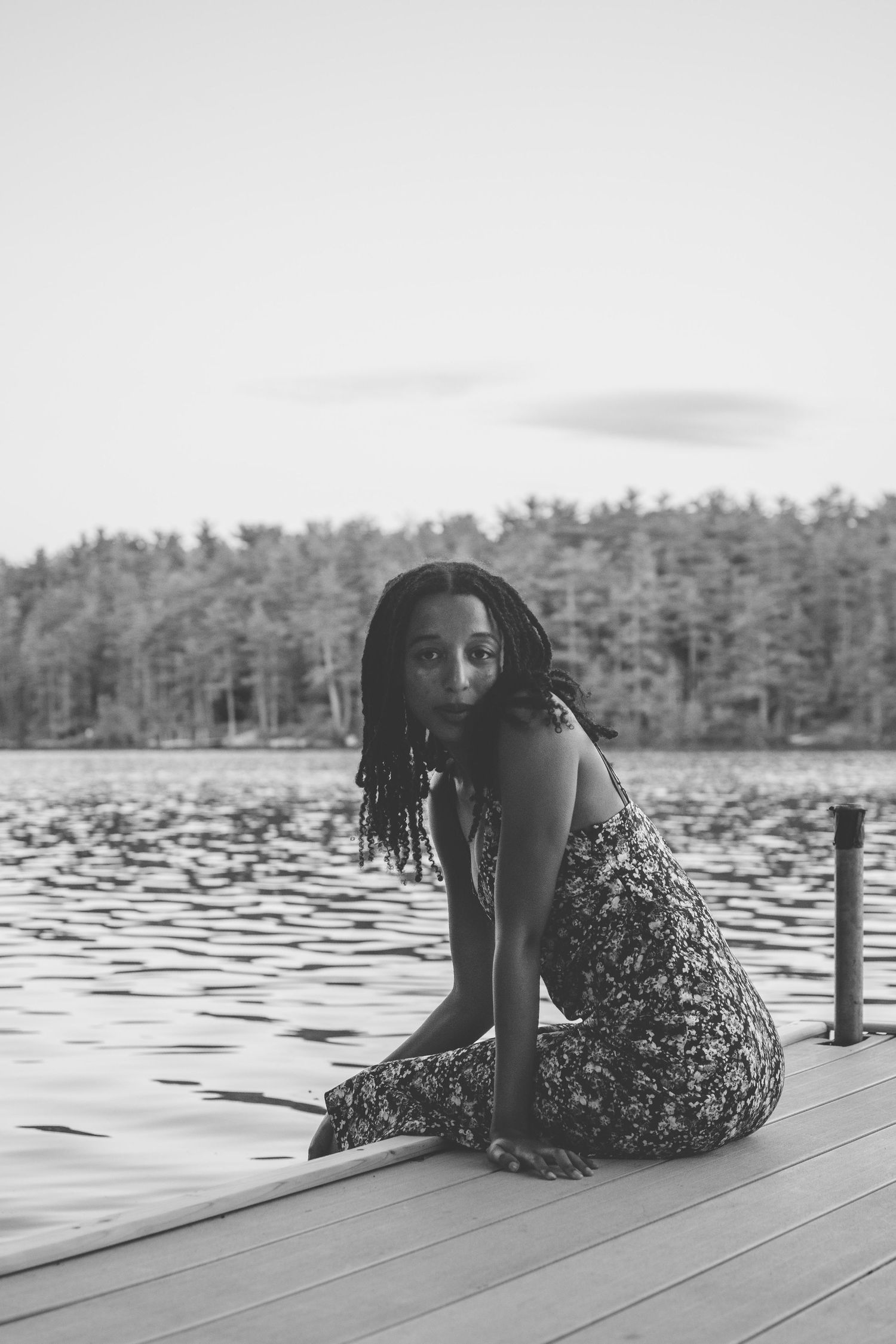 Black and white photo of a young woman with braids sitting on a wooden dock by a serene lake with trees in background.