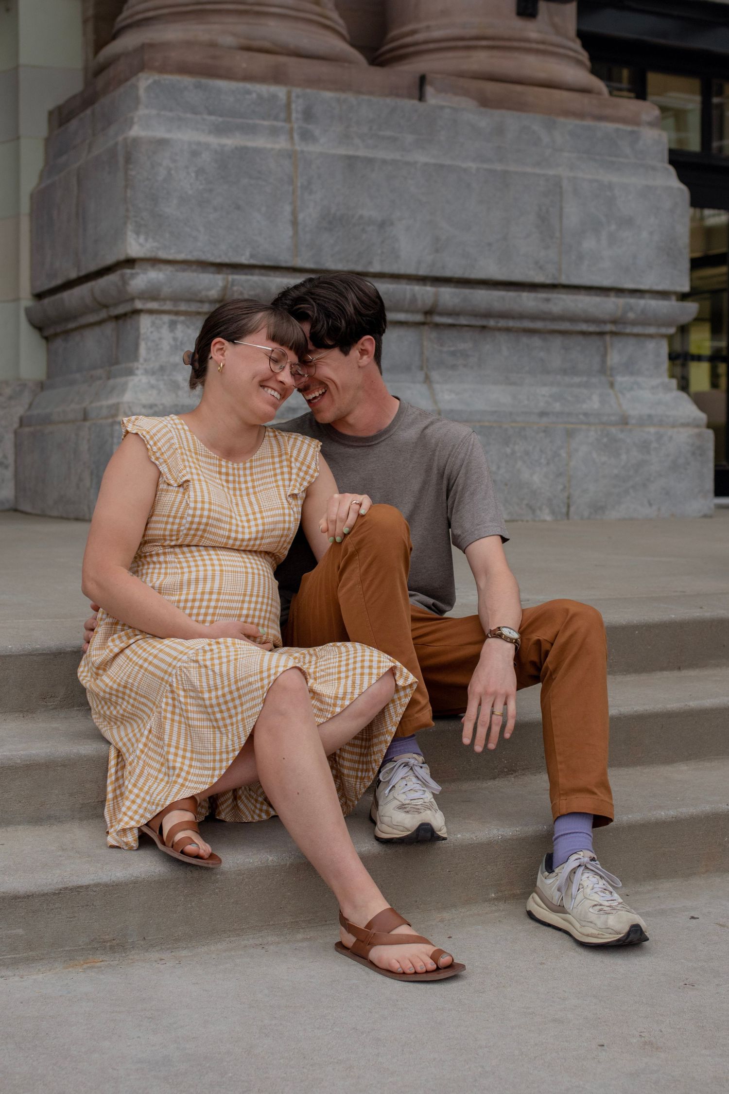 Pregnant woman in yellow dress sits with partner on stone steps, both smiling and sharing a tender moment.