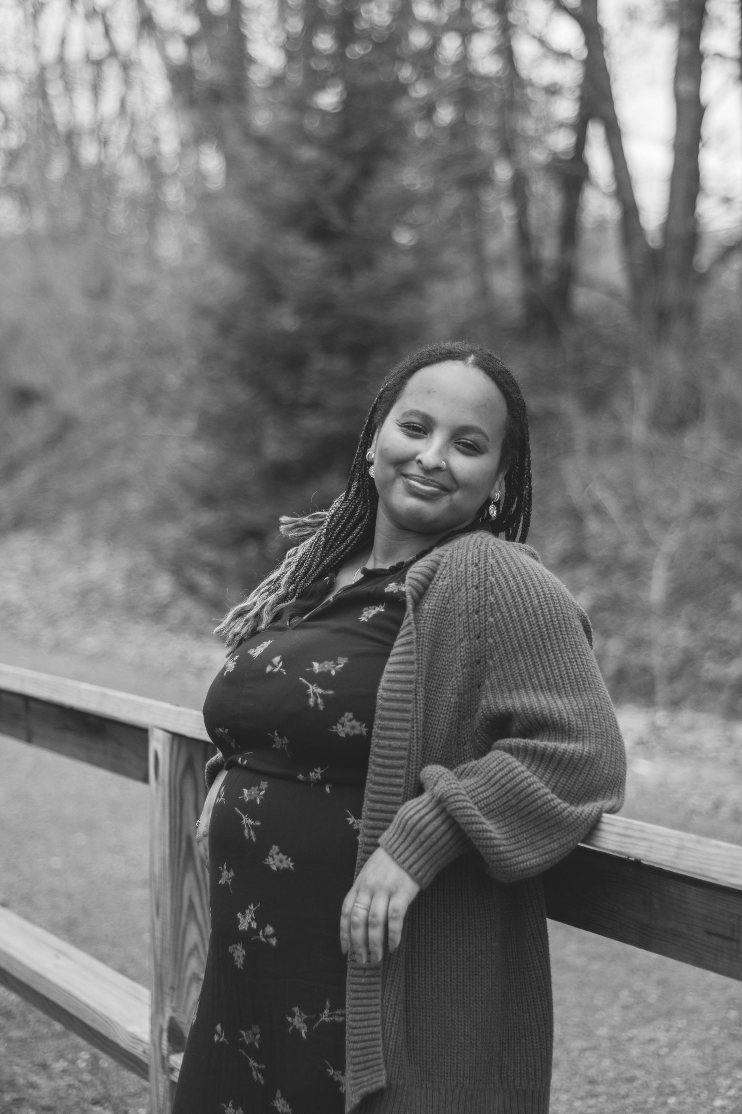 Black and white portrait of a smiling pregnant woman with braids, wearing a floral dress and cardigan, leaning on a wooden fence outdoors.