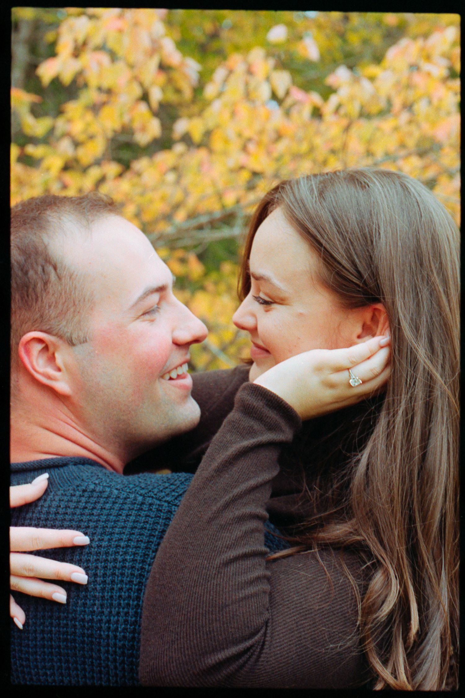 Couple sharing a romantic moment outdoors in autumn, woman wearing engagement ring, colorful fall foliage in background.