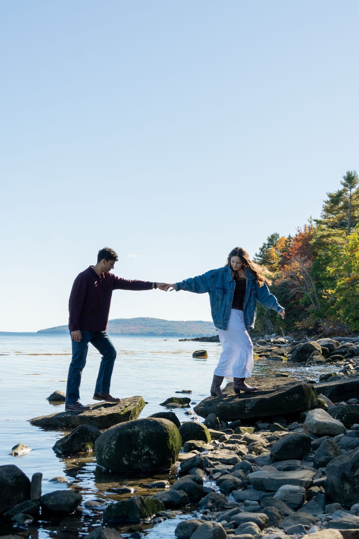 Couple holding hands while balancing on rocky shoreline with calm ocean and autumn trees in background.