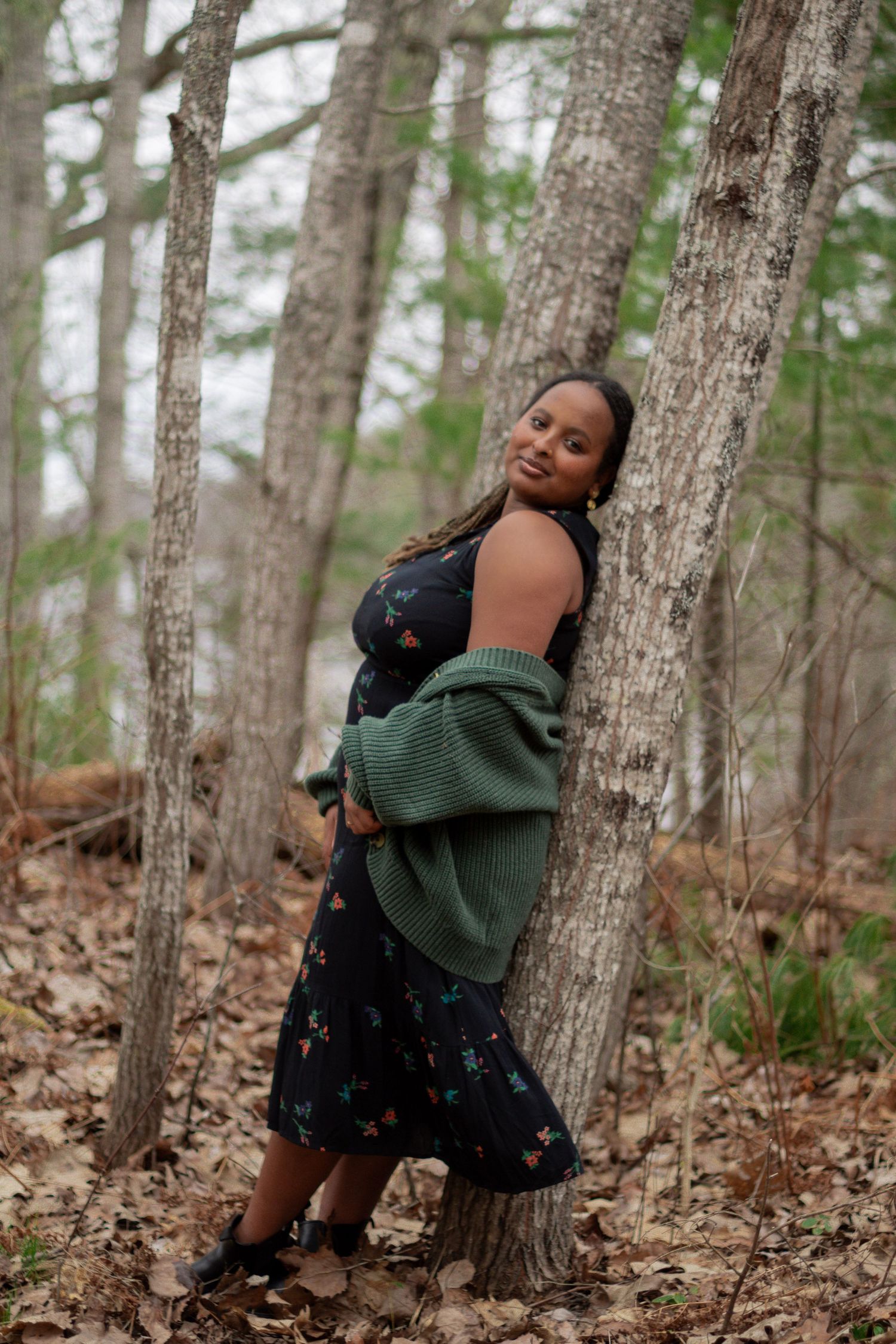 Woman in floral dress and green cardigan leaning against tree in autumn forest setting.