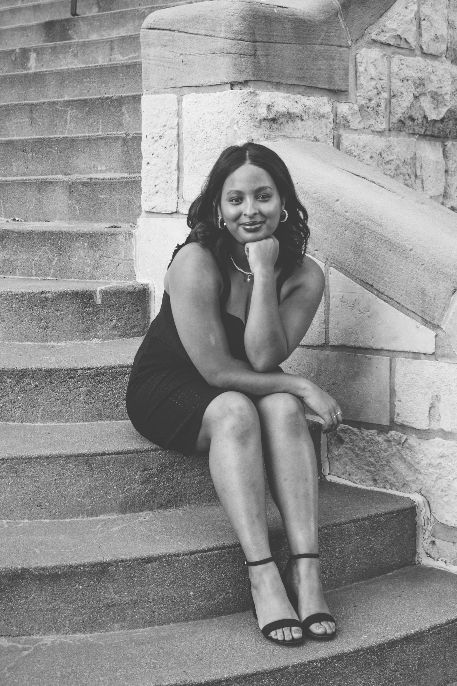 Black and white photo of a young woman in a black dress sitting on stone steps, smiling with hand on chin.