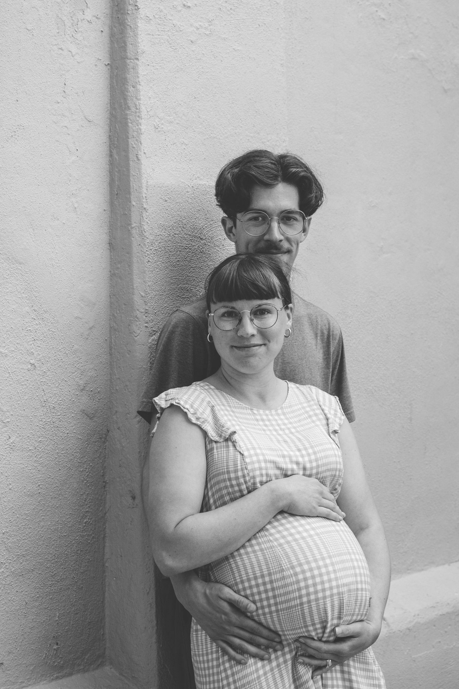 Black and white photo of a pregnant woman in a plaid dress holding her belly, standing with a man against a wall.