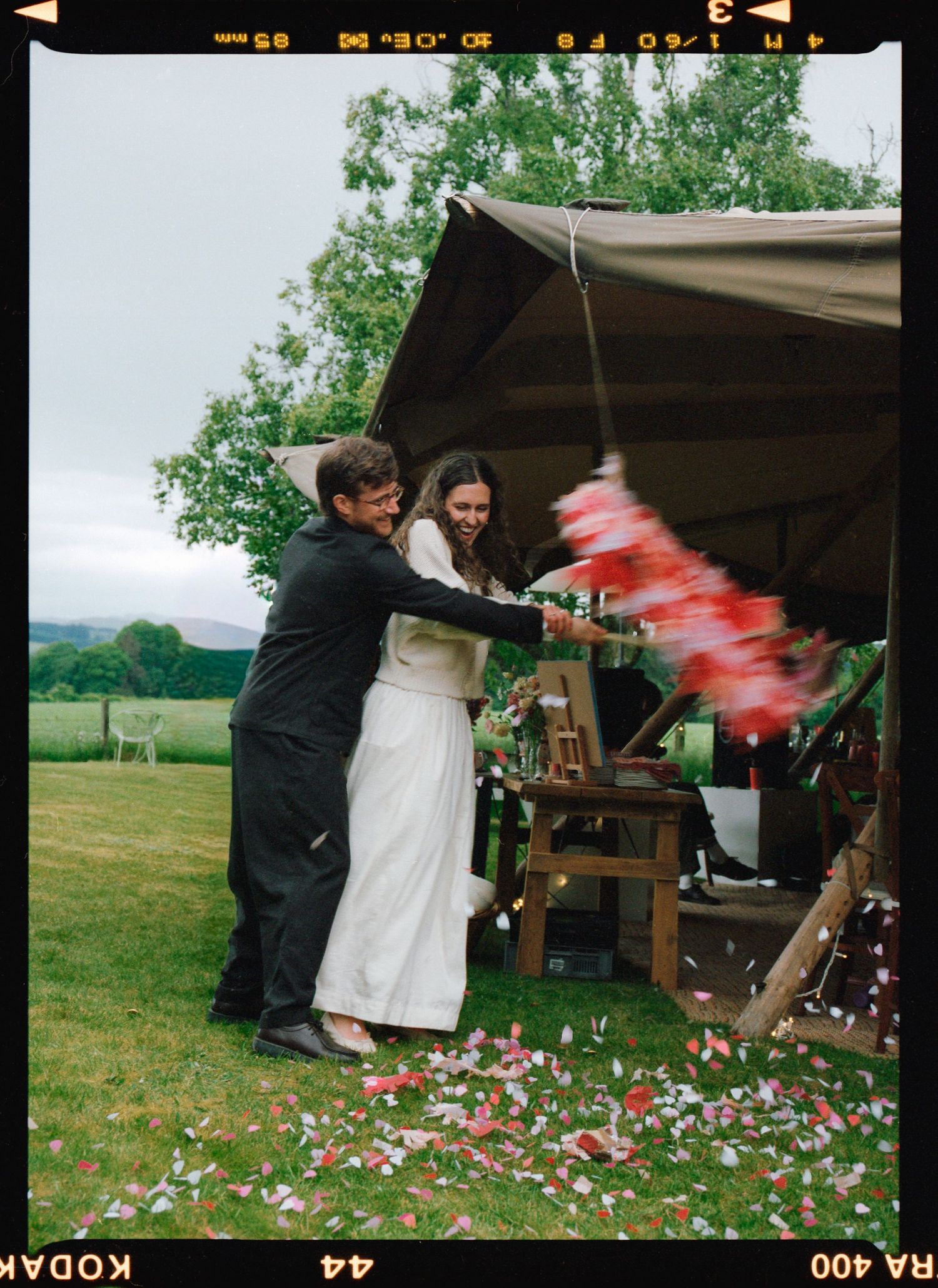 Newlywed couple celebrating outdoors under a tent, tossing red and white confetti on a green lawn.