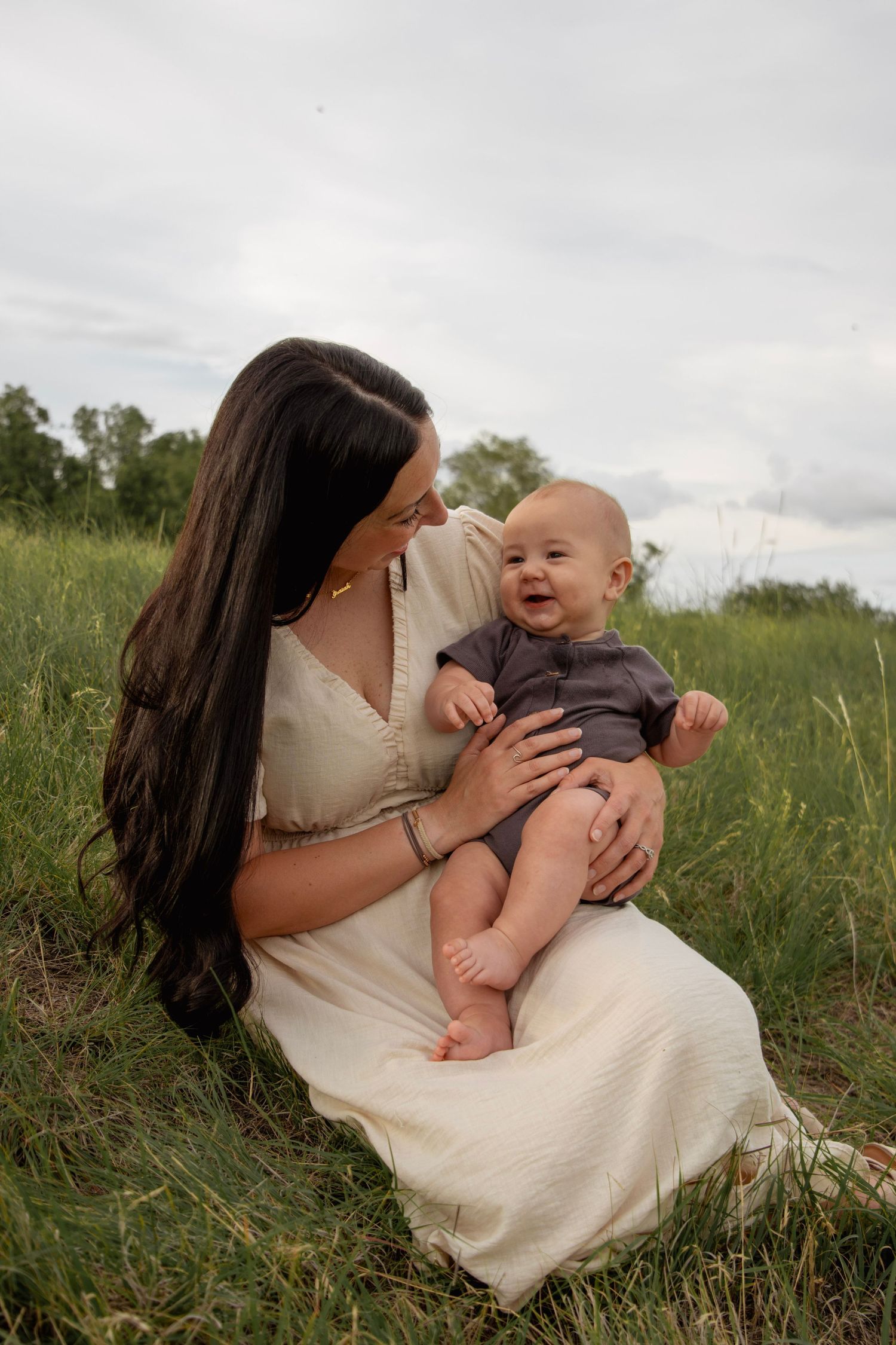 A mother in a white dress sits in a green field holding her smiling baby in a gray outfit.