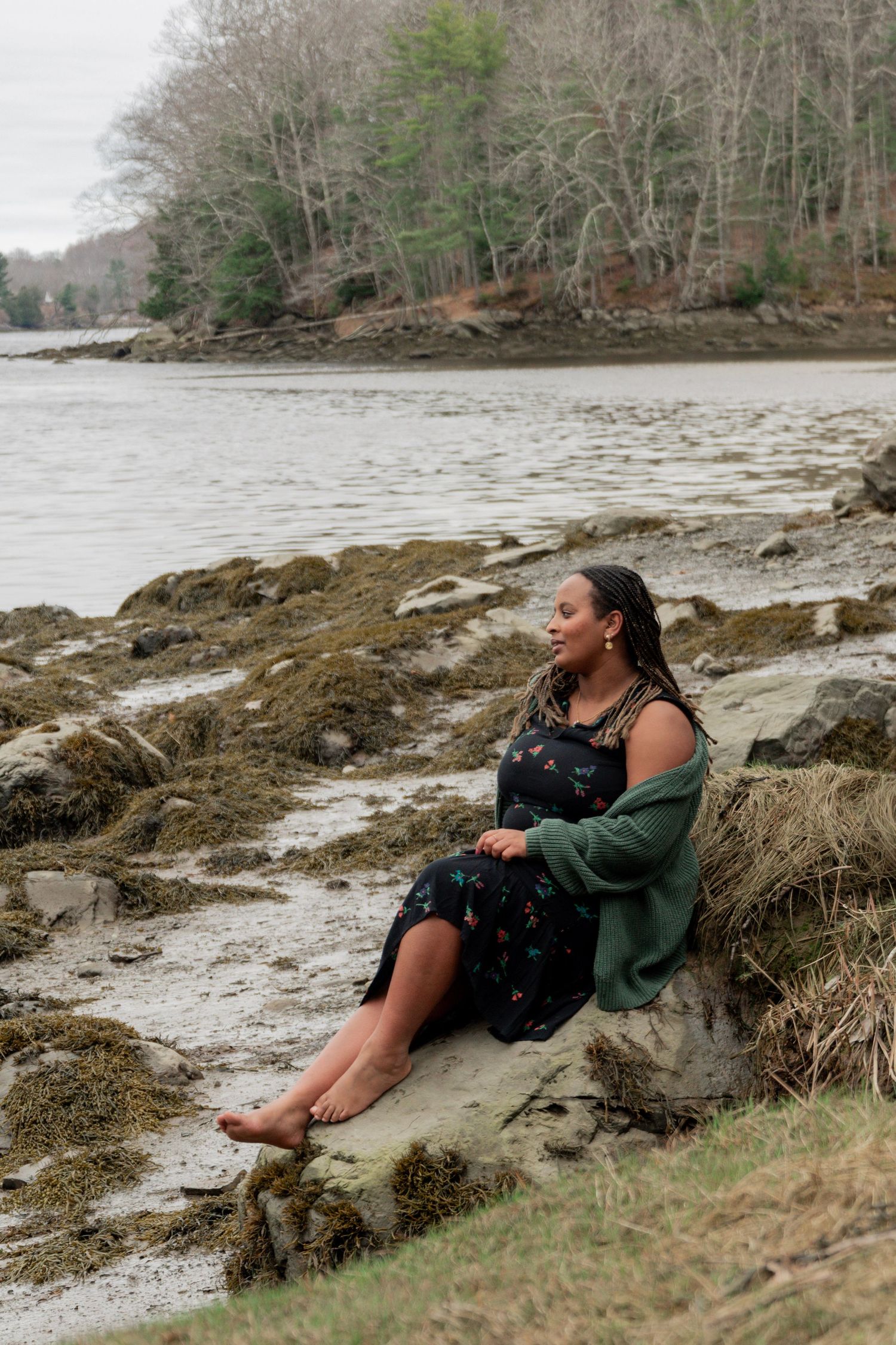 Woman in floral dress and green cardigan sits barefoot on rocky shoreline beside calm water and bare trees.