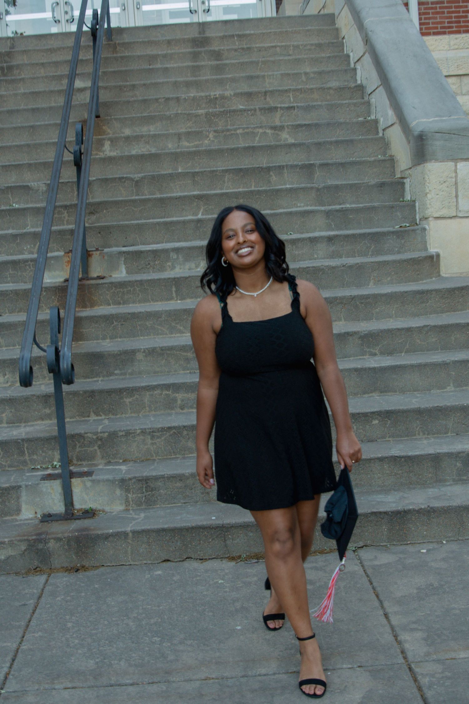 Smiling young woman in black dress holding graduation cap on steps of academic building.