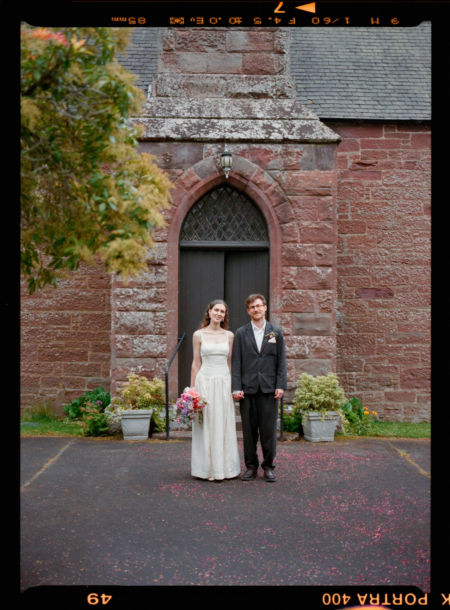 Bride and groom standing hand in hand outside a historic red stone church with arched doorway on their wedding day.
