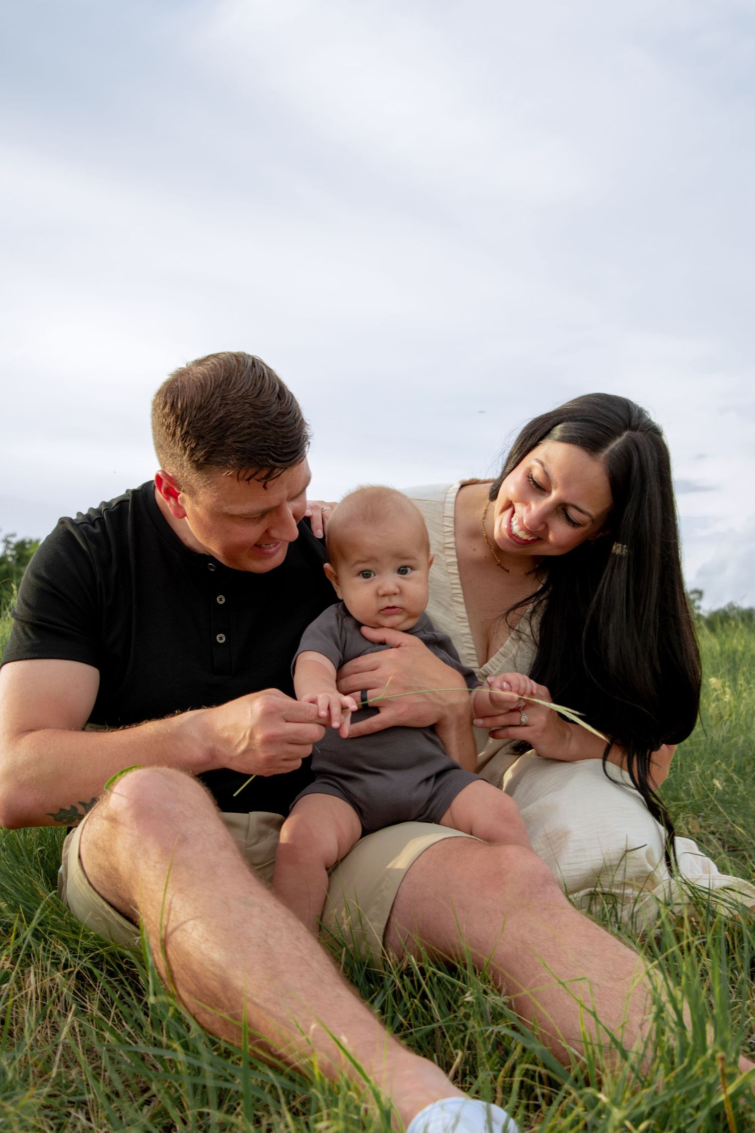 Happy young family with baby sitting on green grass outdoors, parents smiling at infant on cloudy day.