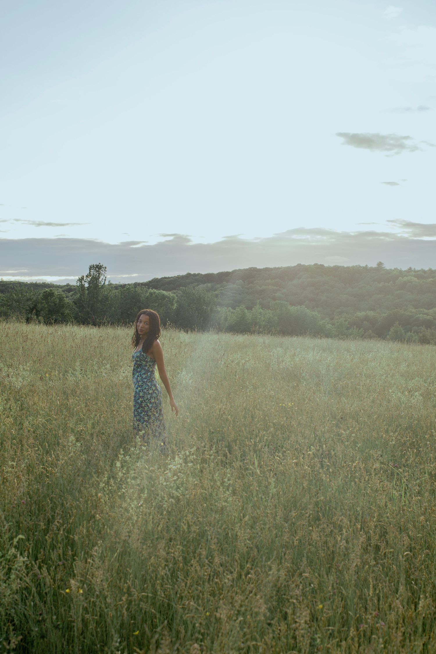 A woman in a floral sundress stands in a lush green meadow with rolling hills and trees in the soft evening light.