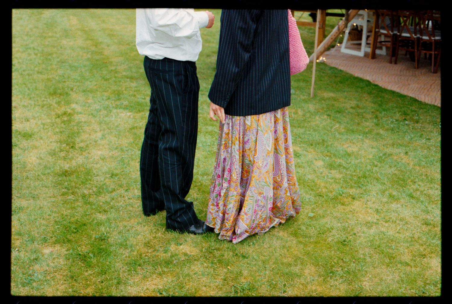 Couple standing on green grass, man in pinstripe trousers, woman in floral maxi skirt, outdoor garden setting.