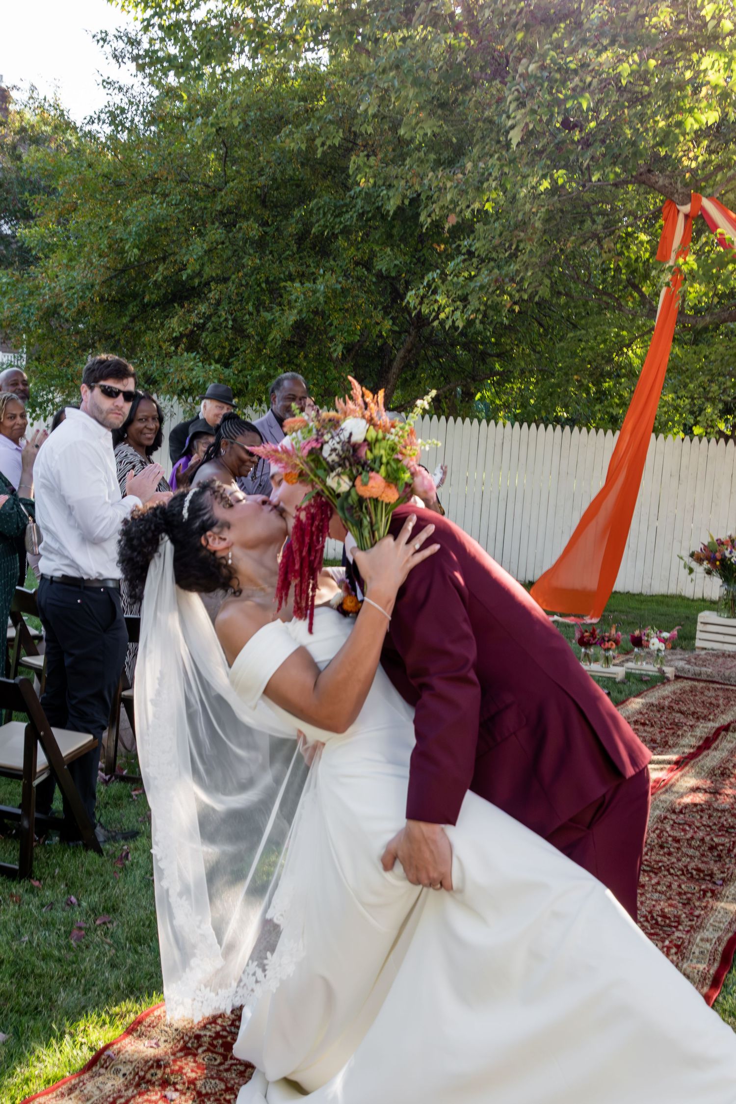 Bride and groom share a romantic dip kiss at outdoor wedding ceremony with guests and white fence backdrop.