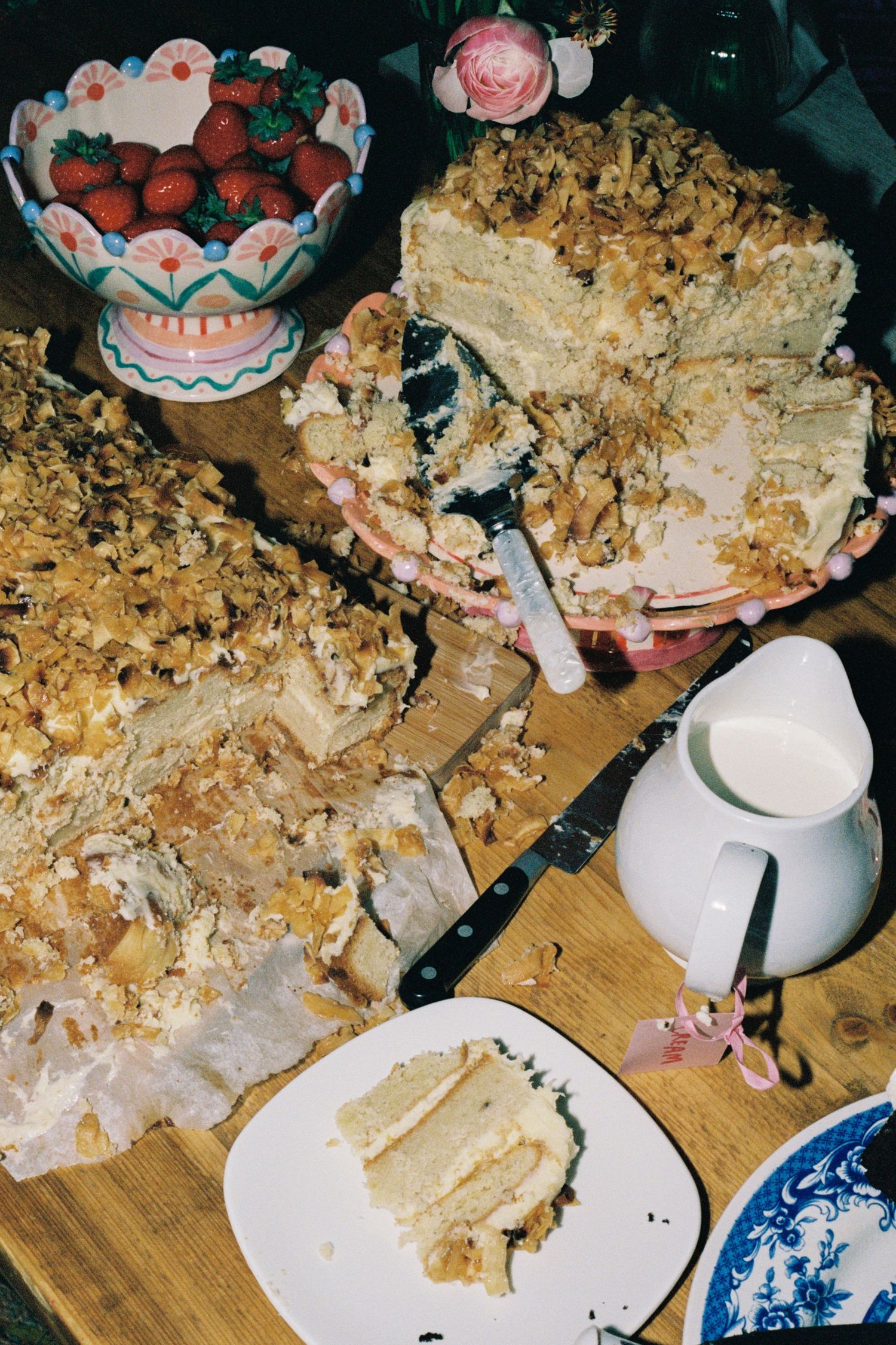 Partially sliced layered cake with walnut topping on wooden board, beside strawberries in decorative bowl and milk pitcher.