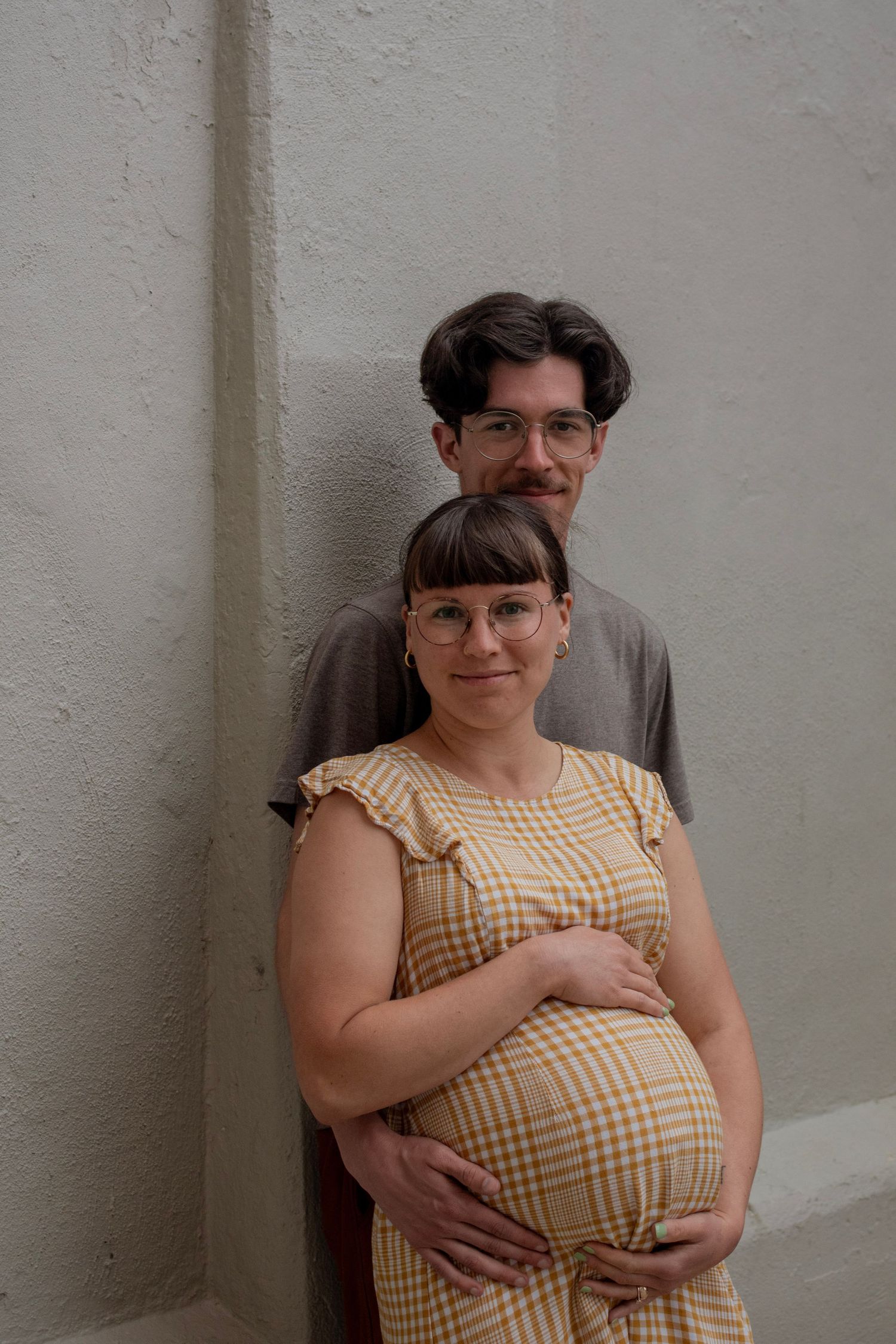 Pregnant woman in plaid dress cradling baby bump with partner standing behind her against a gray wall.