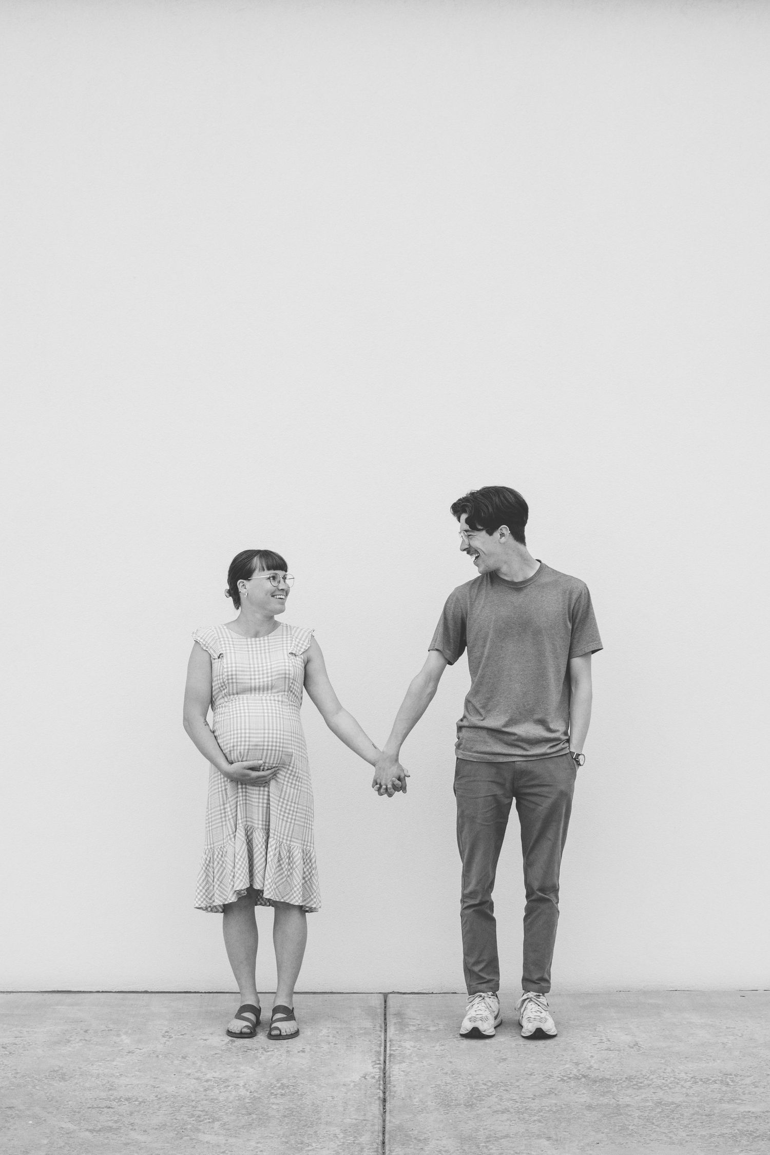 Black and white photo of a smiling pregnant couple holding hands against a white wall.