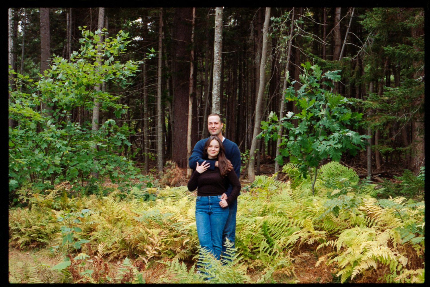 Couple embracing in a lush forest clearing surrounded by ferns and tall evergreen trees.